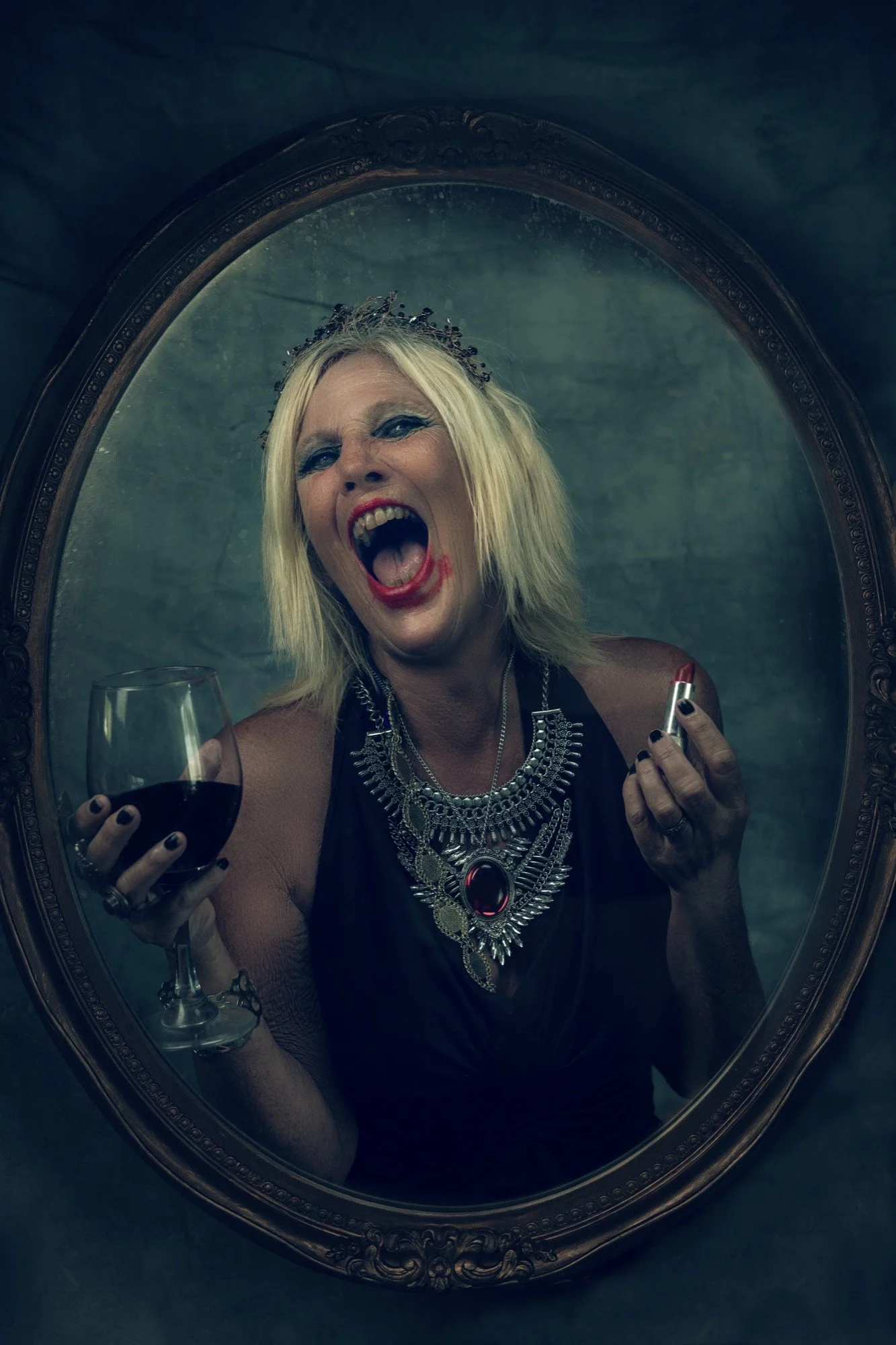 A woman with blonde hair, wearing dark makeup and jewelry, is looking into a mirror and appears to be shouting or singing, holding a glass of red wine in one hand and lipstick in the other.