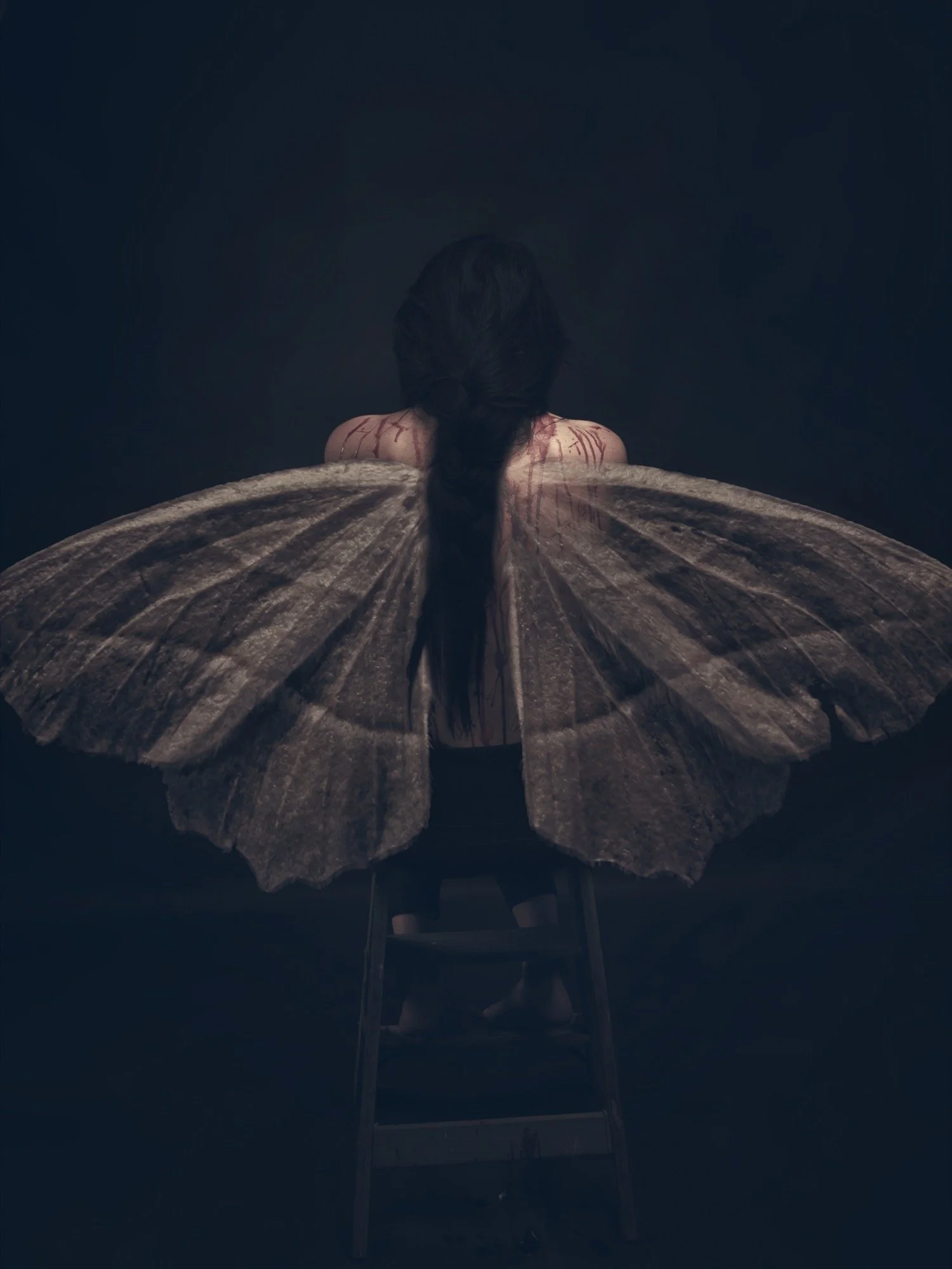 A woman with large butterfly wings sitting on a chair viewed from behind in a dark setting.