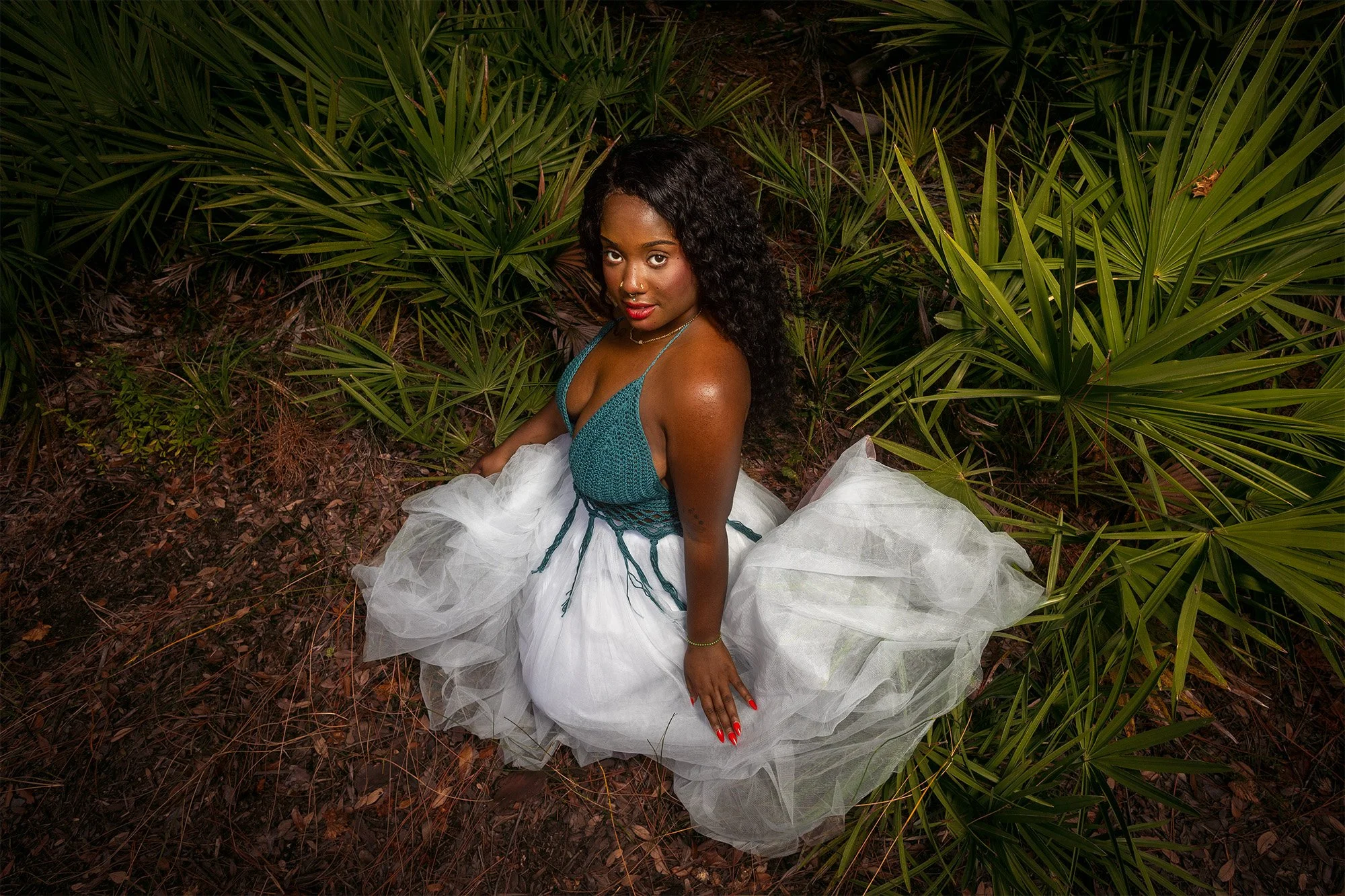 A woman with dark curly hair and medium skin tone wearing a teal top with a white tulle skirt, sitting in a leafy outdoor area with green plants.