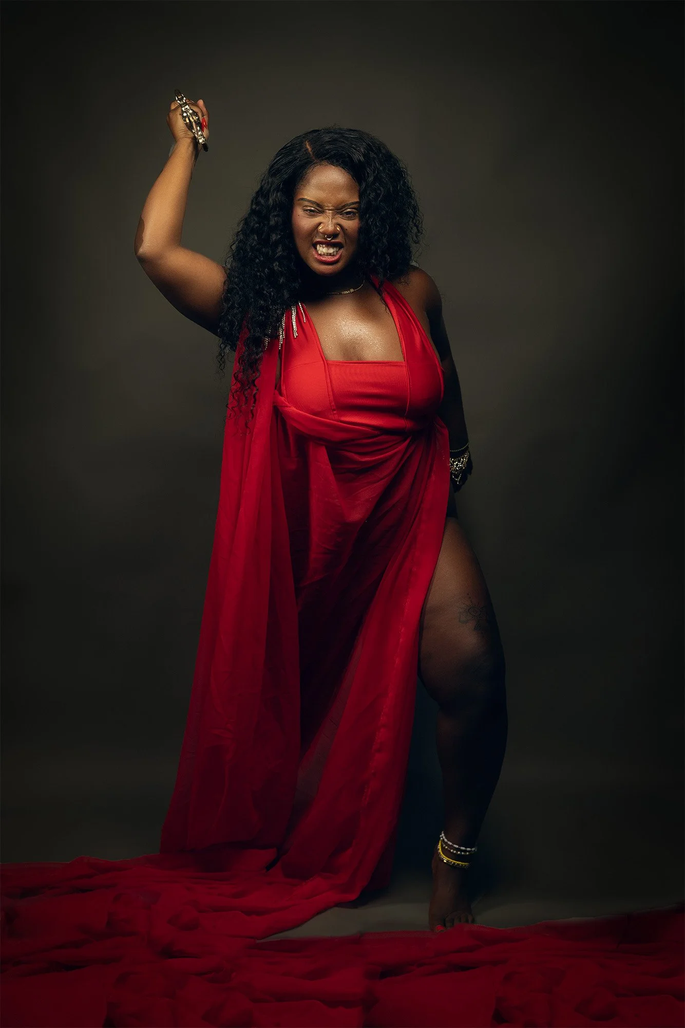 A woman with curly black hair, wearing a red dress with a sheer cape, standing on a dark background. She has a fierce expression, showing her teeth, and is holding an object in her right hand above her head.