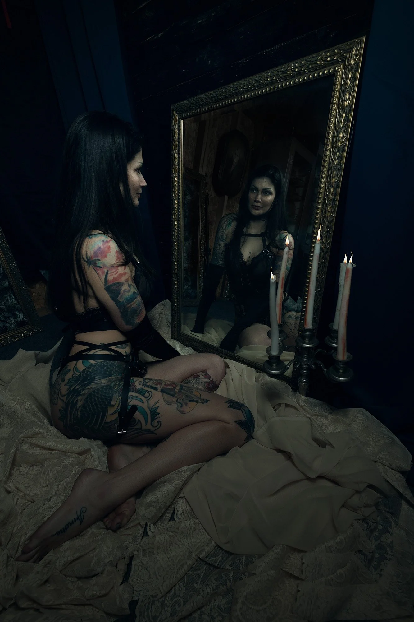 A woman with dark hair and tattoos, wearing black lingerie and stockings, seated on a bed with beige sheets, looking at her reflection in a large ornate mirror surrounded by lit candles in candlesticks.