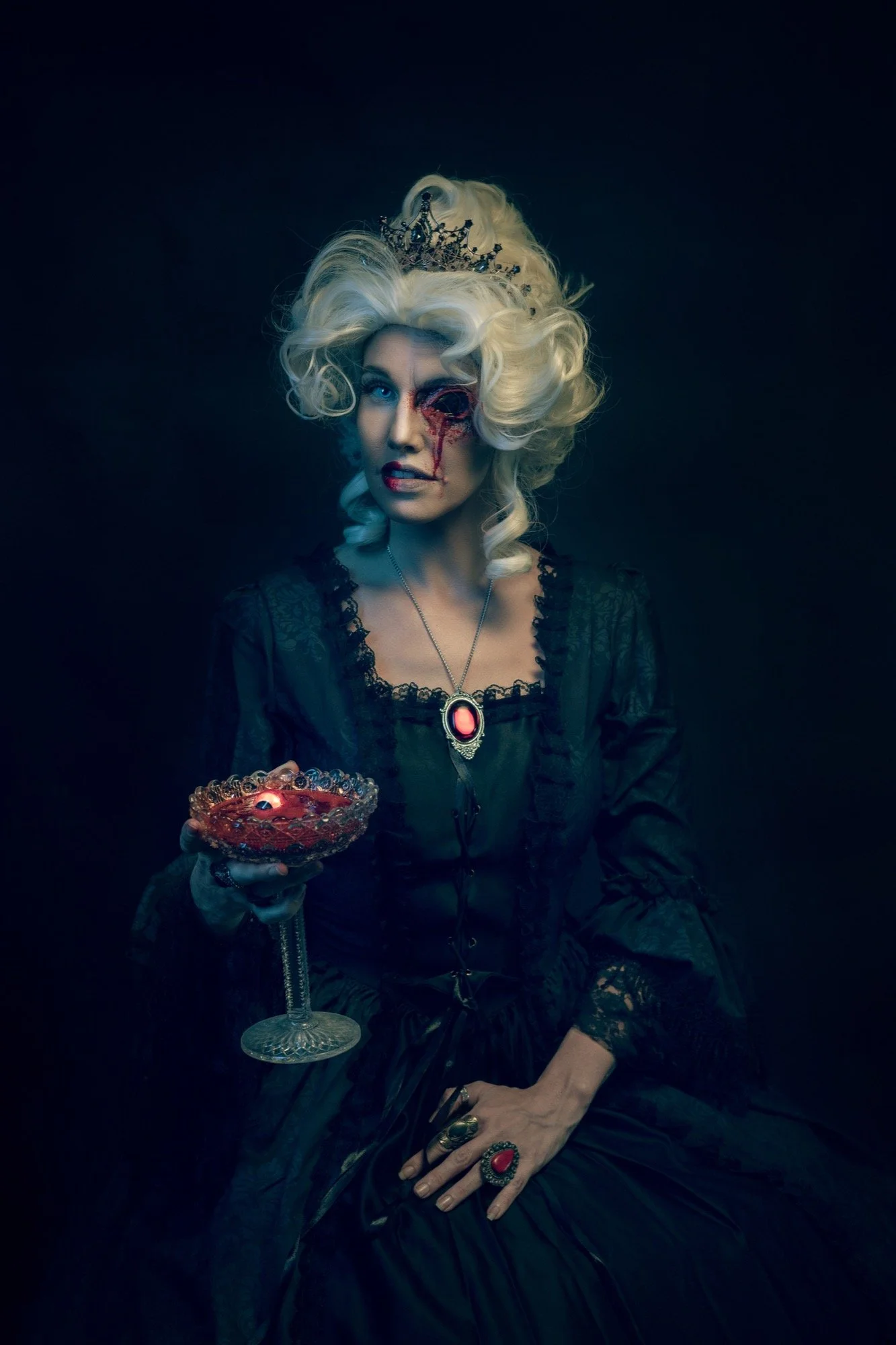 Woman with platinum blonde, curly hair wearing a crown, dressed in black, with blood and makeup on her face, holding a glass bowl with red liquid, seated against a dark background.