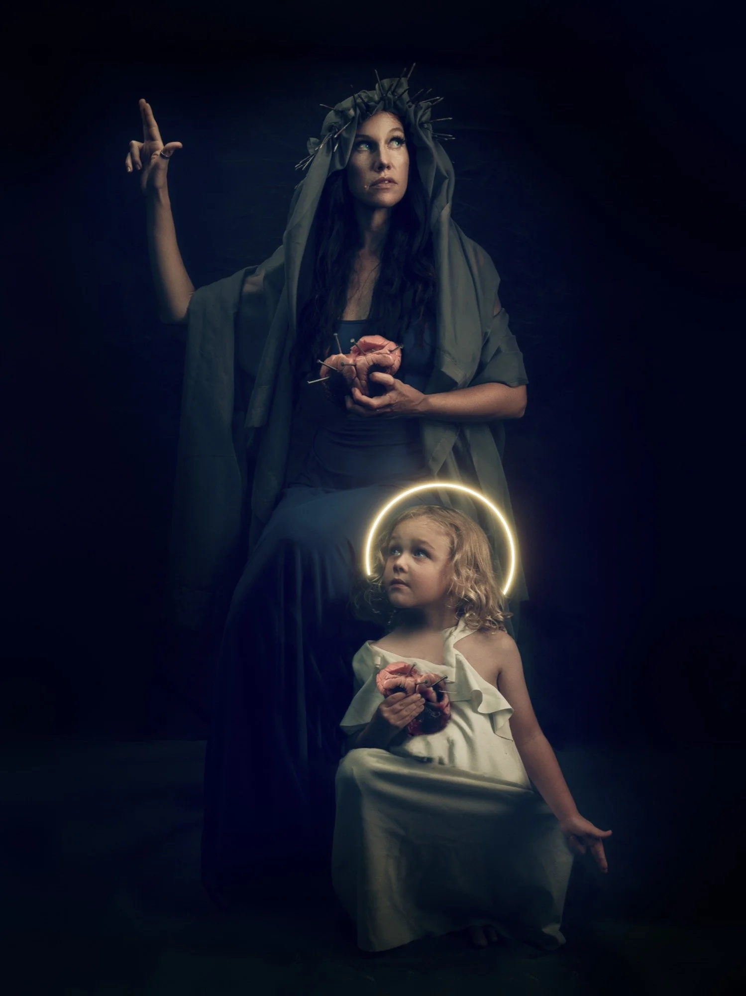 A woman dressed as a medieval or religious figure with a halo and holding a heart, standing behind a young girl with a halo, who is also holding a heart. The scene is dark and dramatic.