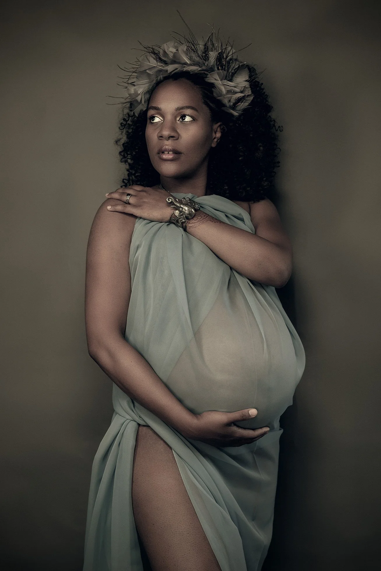 A pregnant woman with curly hair and a feathered headpiece, wearing a sheer, draped fabric, standing against a plain wall, touching her shoulder and holding her belly.