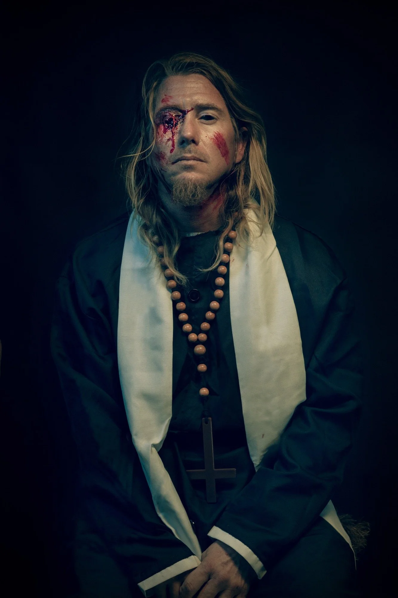 A man dressed as a priest with a cross necklace, sitting against a dark background, with bloody makeup on his face, including a wound around his eye and scratches on his face.