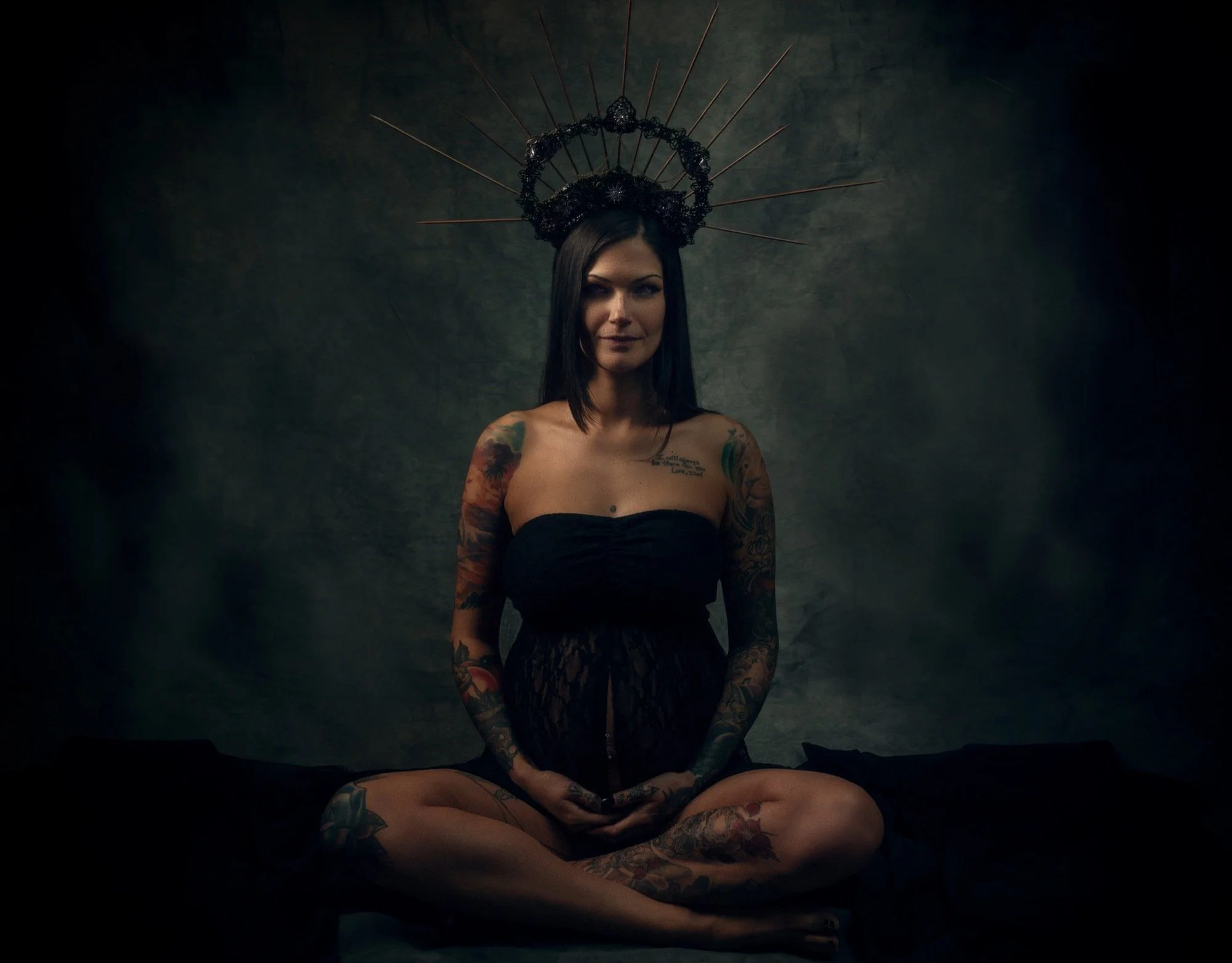 A woman with long dark hair and tattoos, sitting cross-legged with hands in lap, wearing a strapless black top and lace skirt, sits against a dark textured background. She wears an ornate halo-like headpiece with spikes and circular elements.