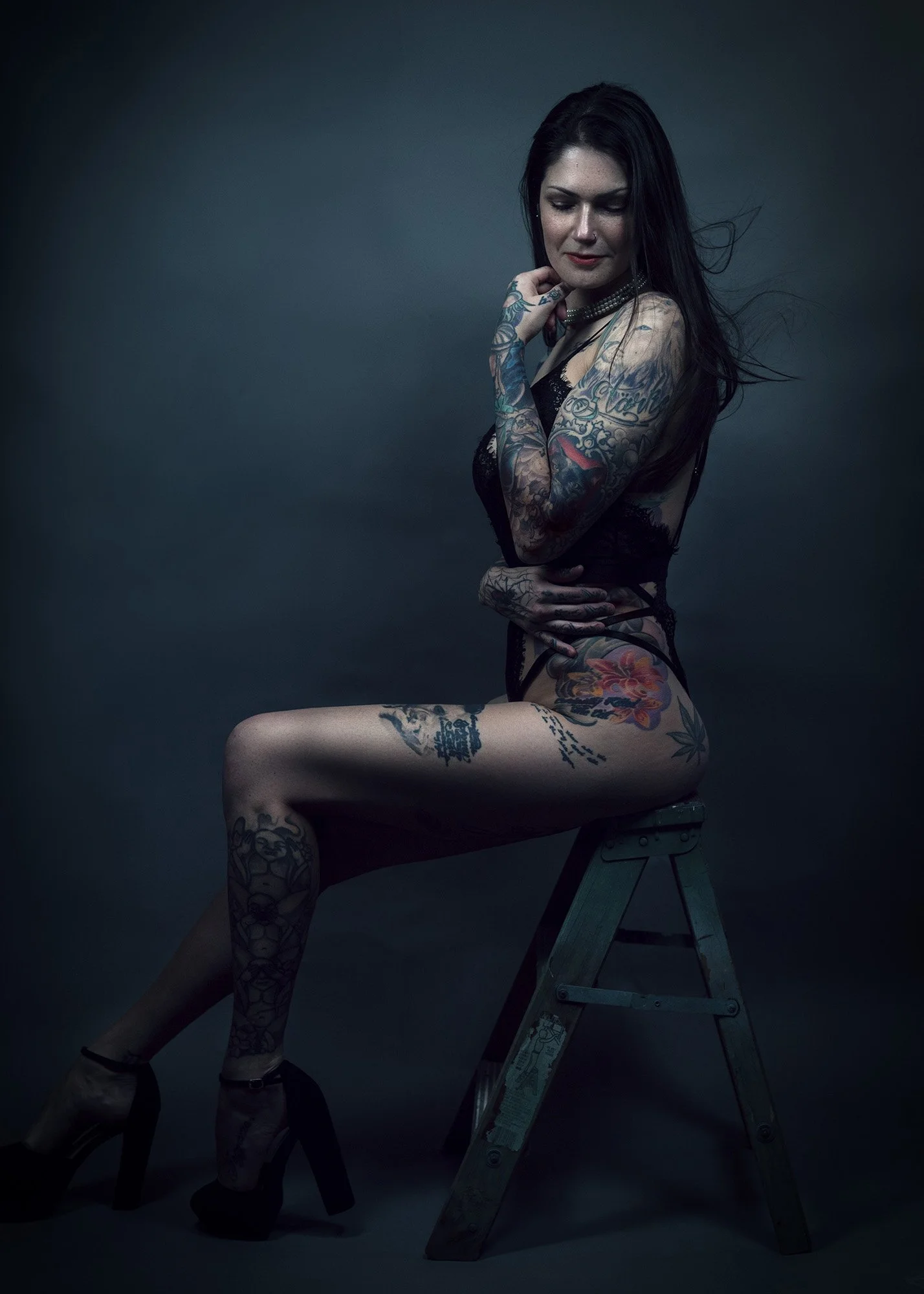 A tattooed woman with dark hair sitting on a ladder in a dark studio, wearing black lingerie and high heels, with a contemplative expression.
