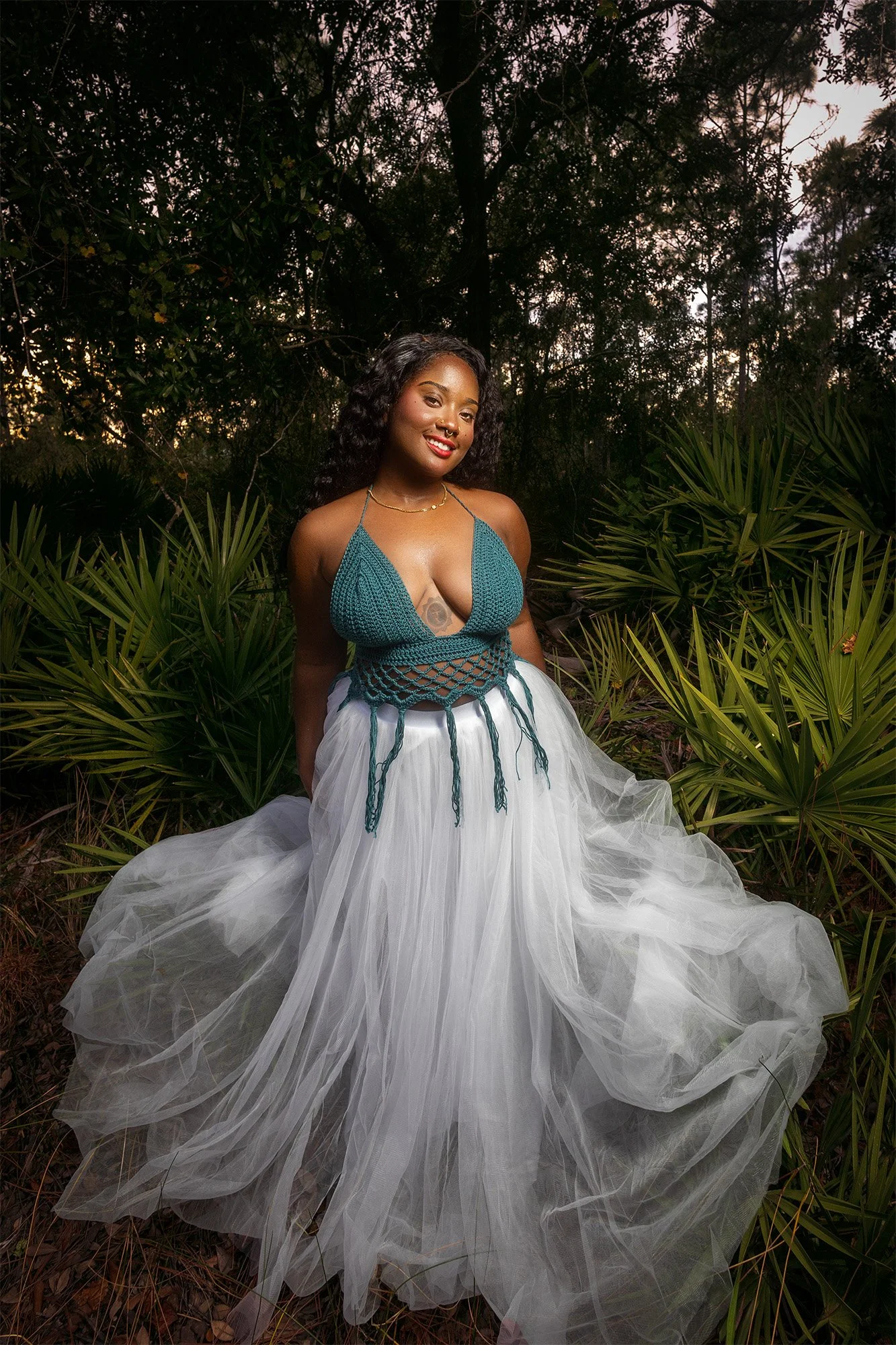 A woman standing in a lush, green forest at sunset, wearing a teal crochet top with a deep neckline and a flowing white tulle skirt.