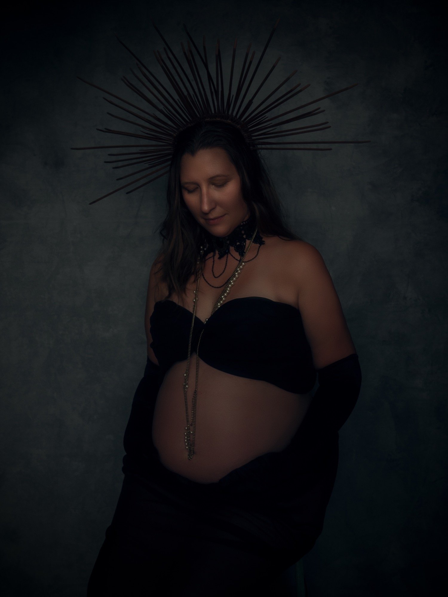 A pregnant woman with long dark hair wearing black gloves, a black strapless top, and layered necklaces, standing against a dark textured background, with a silver spiked halo around her head.