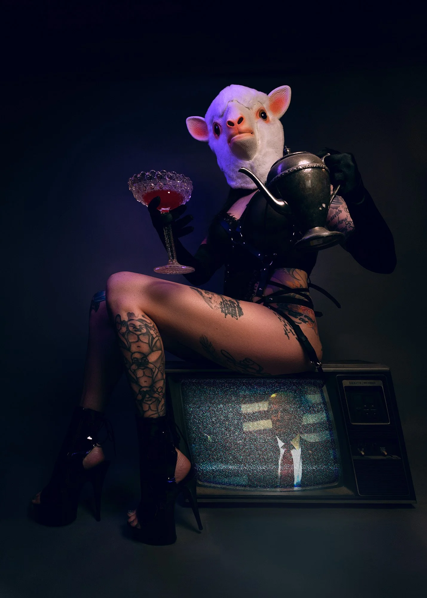 Person with a sheep mask sitting on a vintage television set, wearing black high heels, revealing tattoos, holding a glass of red drink in one hand and a teapot in the other, with a TV screen showing a man in a suit.