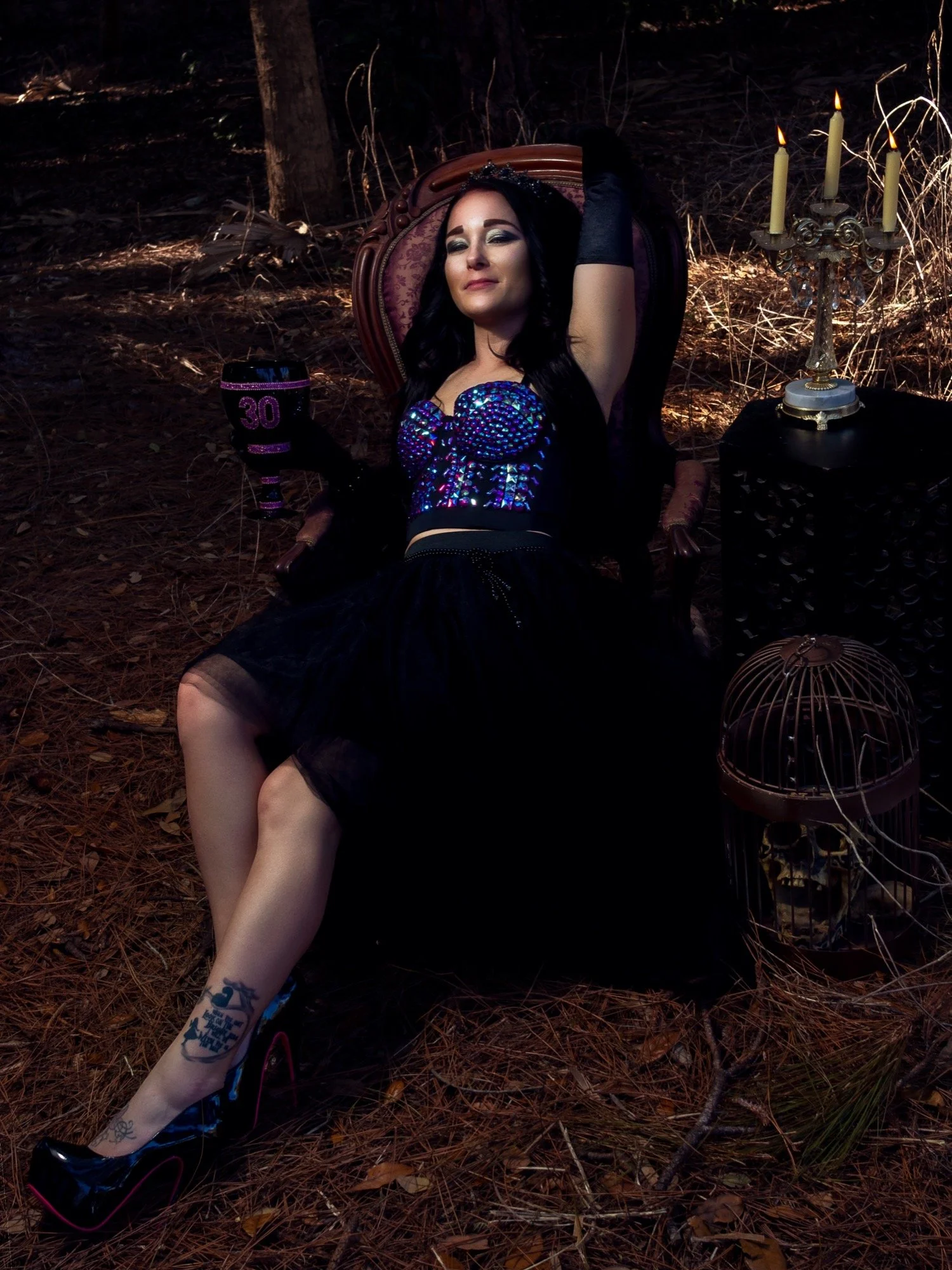 Woman dressed in dark, glamorous attire, sitting in a vintage armchair outdoors at night, holding a black cup with the number 30, surrounded by candles and decorative objects.
