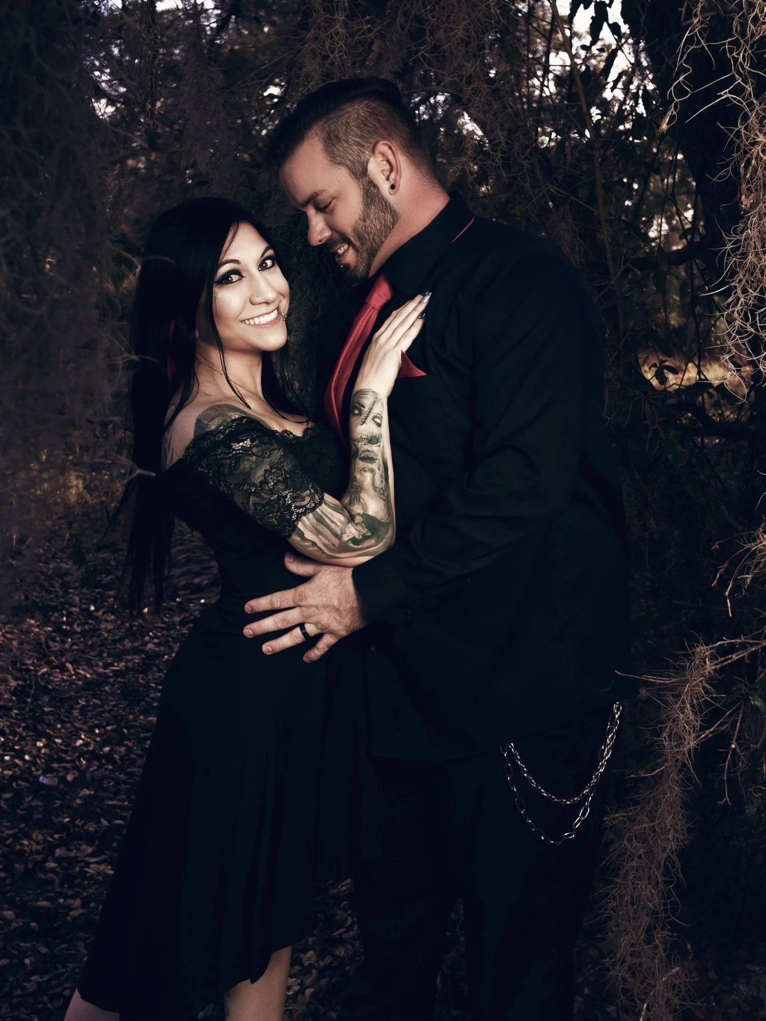 A smiling woman with dark hair and tattoos on her arm, and a man with a beard, dark hair, and earrings, dressed in black with a red tie, embrace each other in a wooded area at dusk.