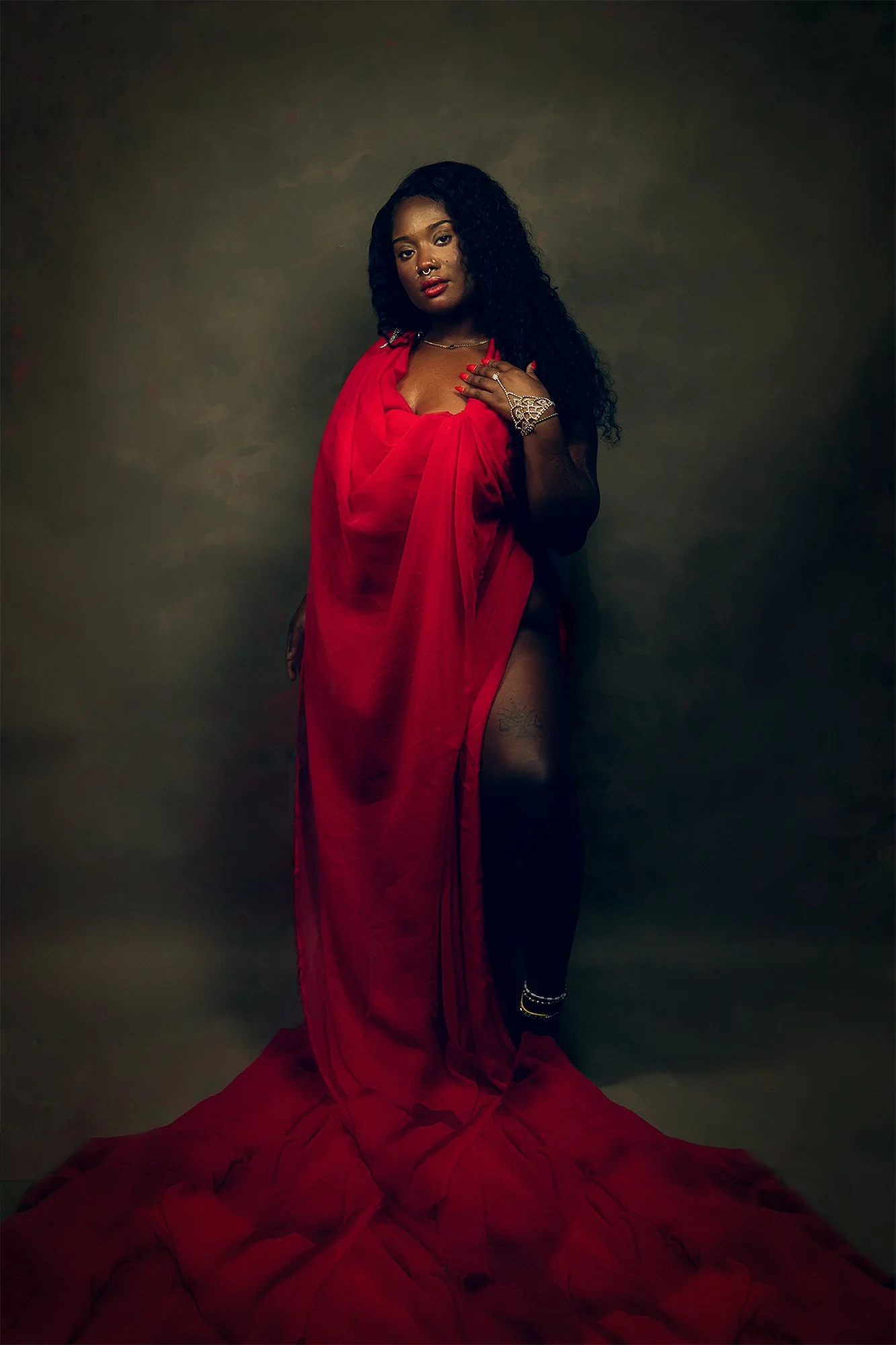 A woman with dark curly hair wearing a red sheer dress, standing against a dark background, with one hand on her chest and the other by her side, accessorized with jewelry