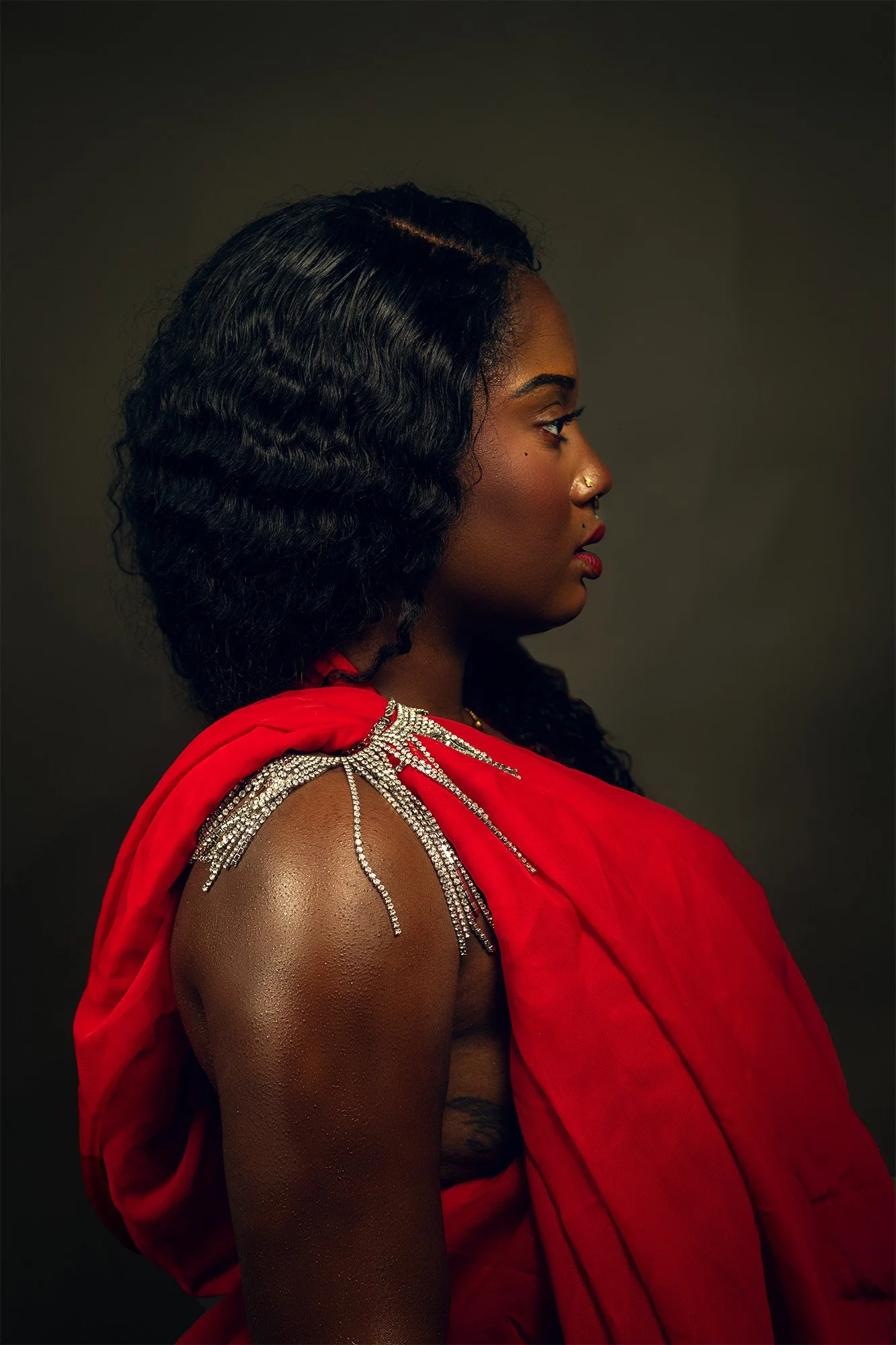 A side profile of a woman with dark wavy hair, dressed in a red garment with decorative silver beaded shoulder embellishments, set against a dark background.