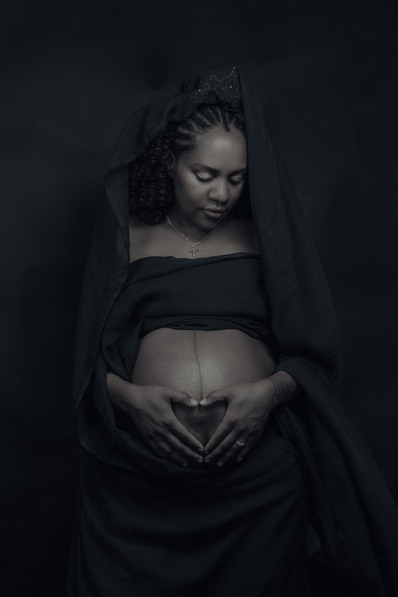 A pregnant woman dressed in black with a black veil, holding her belly in a heart shape with her hands. She is wearing a cross necklace and has dark curly hair. The background is black.