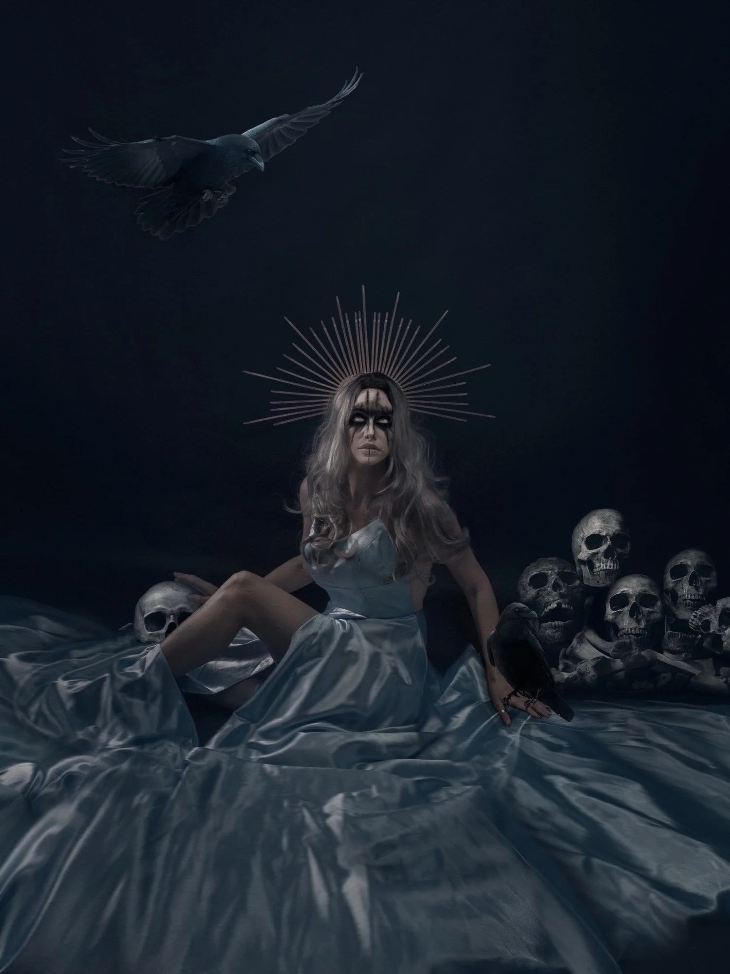 A woman with long gray hair and dark makeup, wearing a white dress, sitting on flowing fabric surrounded by skulls and black birds, with a dark background and a halo-like headpiece behind her head.