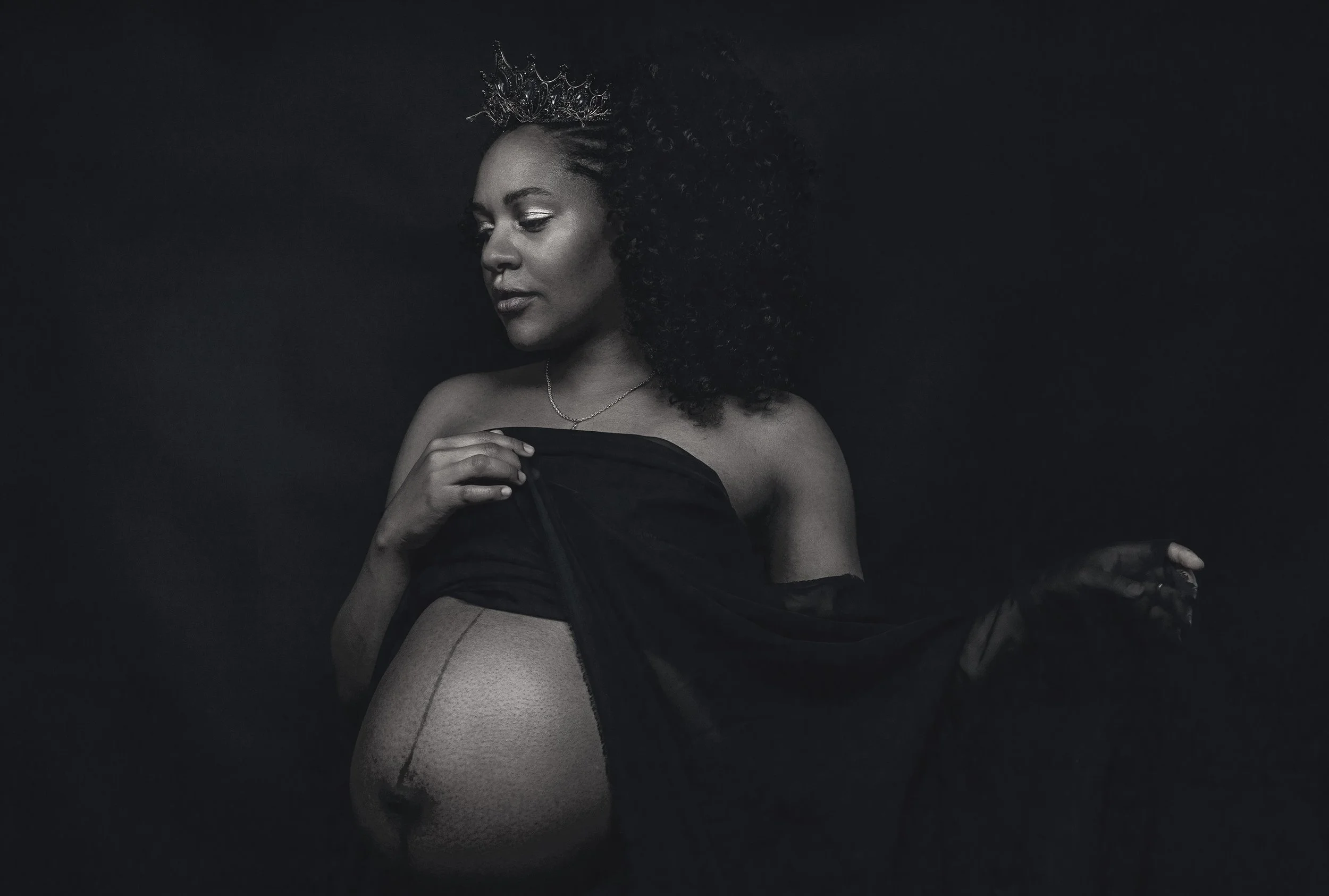 Black and white portrait of a pregnant woman with curly hair, wearing a tiara and a necklace, holding a sheer black fabric.