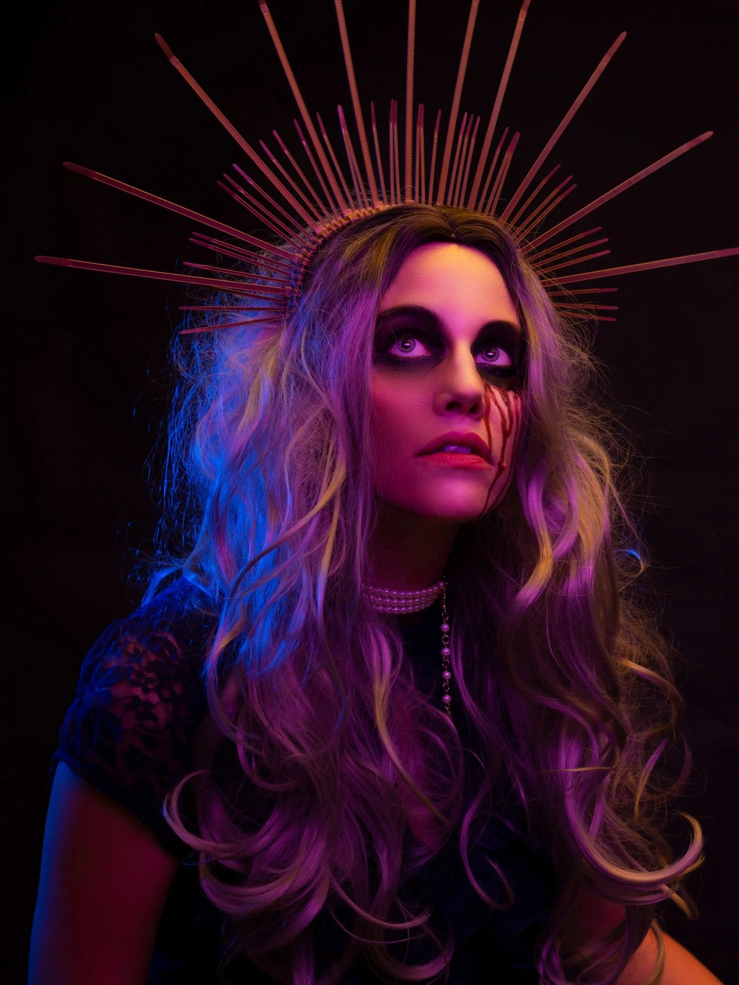 A woman with long, curly hair illuminated in purple and blue lights. She wears a headpiece resembling a halo with radiating lines, dark eye makeup with black and purple, and a pearl necklace. The background is black.