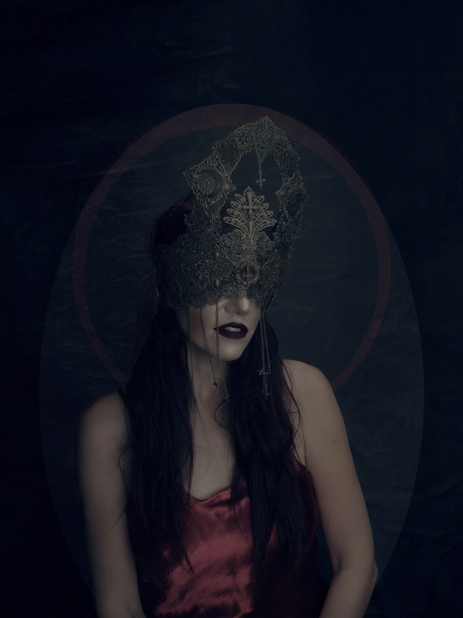 Woman with long dark hair wearing a dark red satin top, ornate metallic mask covering her eyes, dark lipstick, and cross earrings, against a dark background with a faint circular glow.