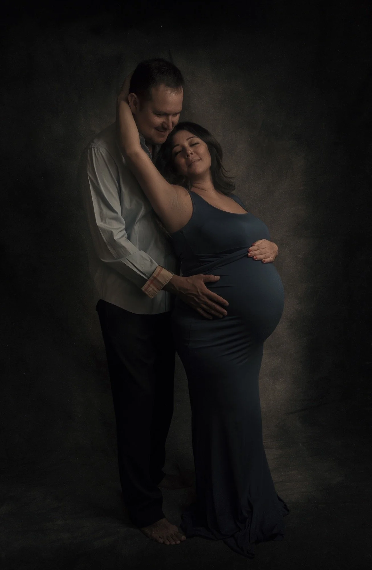 A pregnant woman and a man embracing and smiling, with the woman touching her belly, against a dark background.