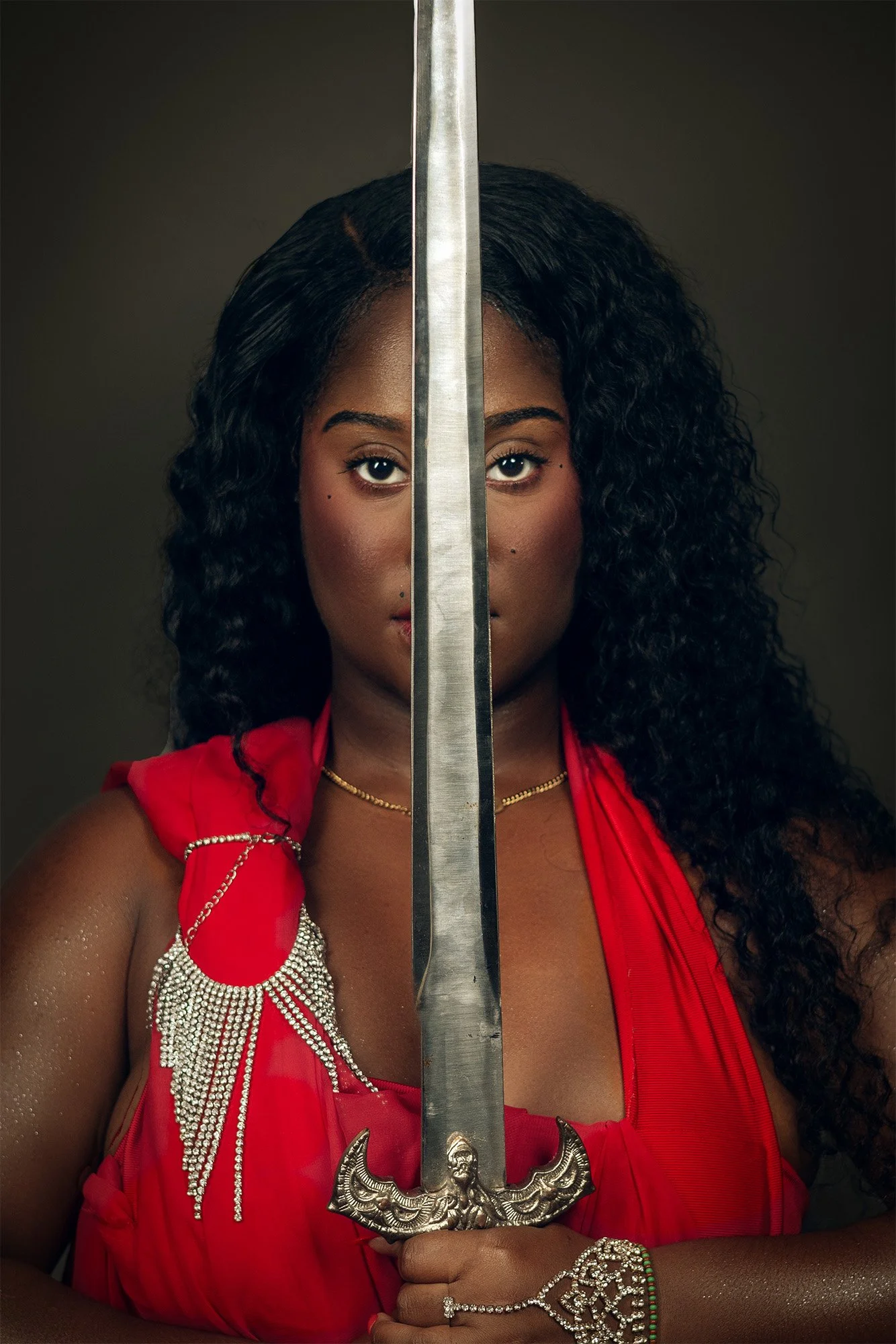 A woman with dark, curly hair and dark eyes holds a sword vertically in front of her face, obscuring the nose and mouth. She is wearing a red dress with decorative silver chains and jewelry, including a bracelet and a necklace. The background is dark