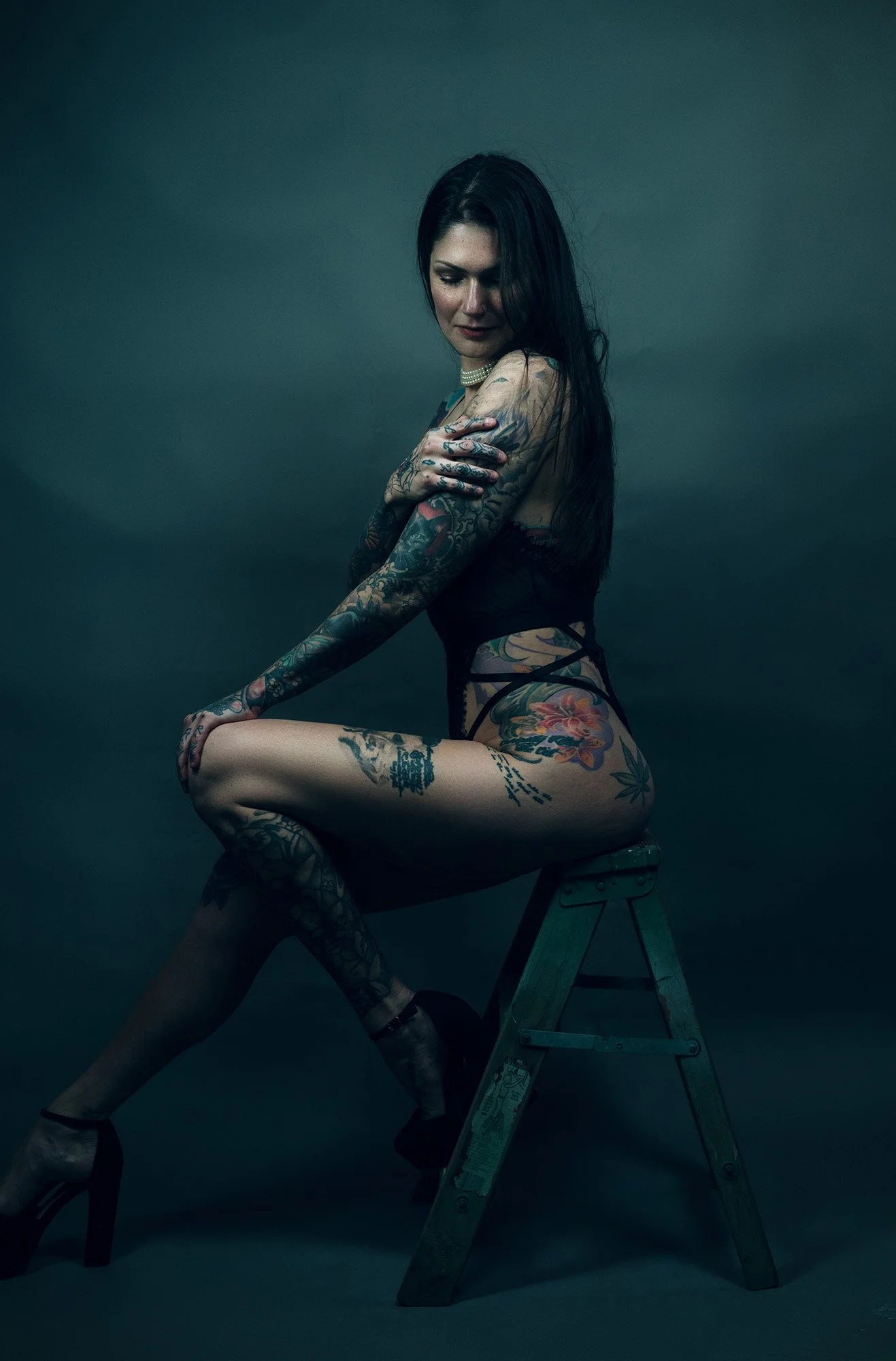 A woman with long dark hair and tattoos, wearing black lingerie and high heels, seated on a step stool in a dark studio setting.