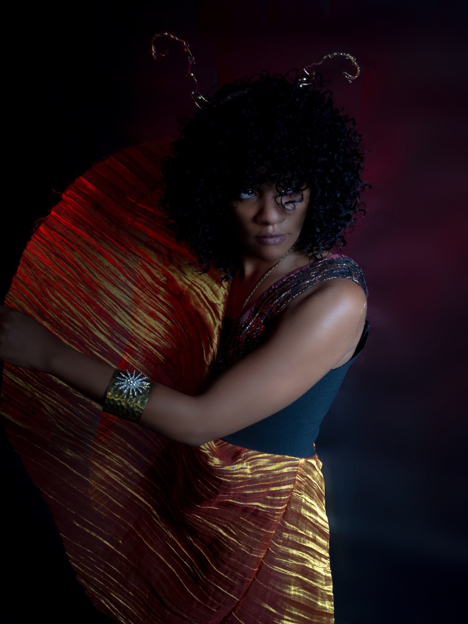 Woman with curly black hair wearing a black top, gold bracelet, and a colorful red and gold fabric, against a dark background with pink and purple light accents.