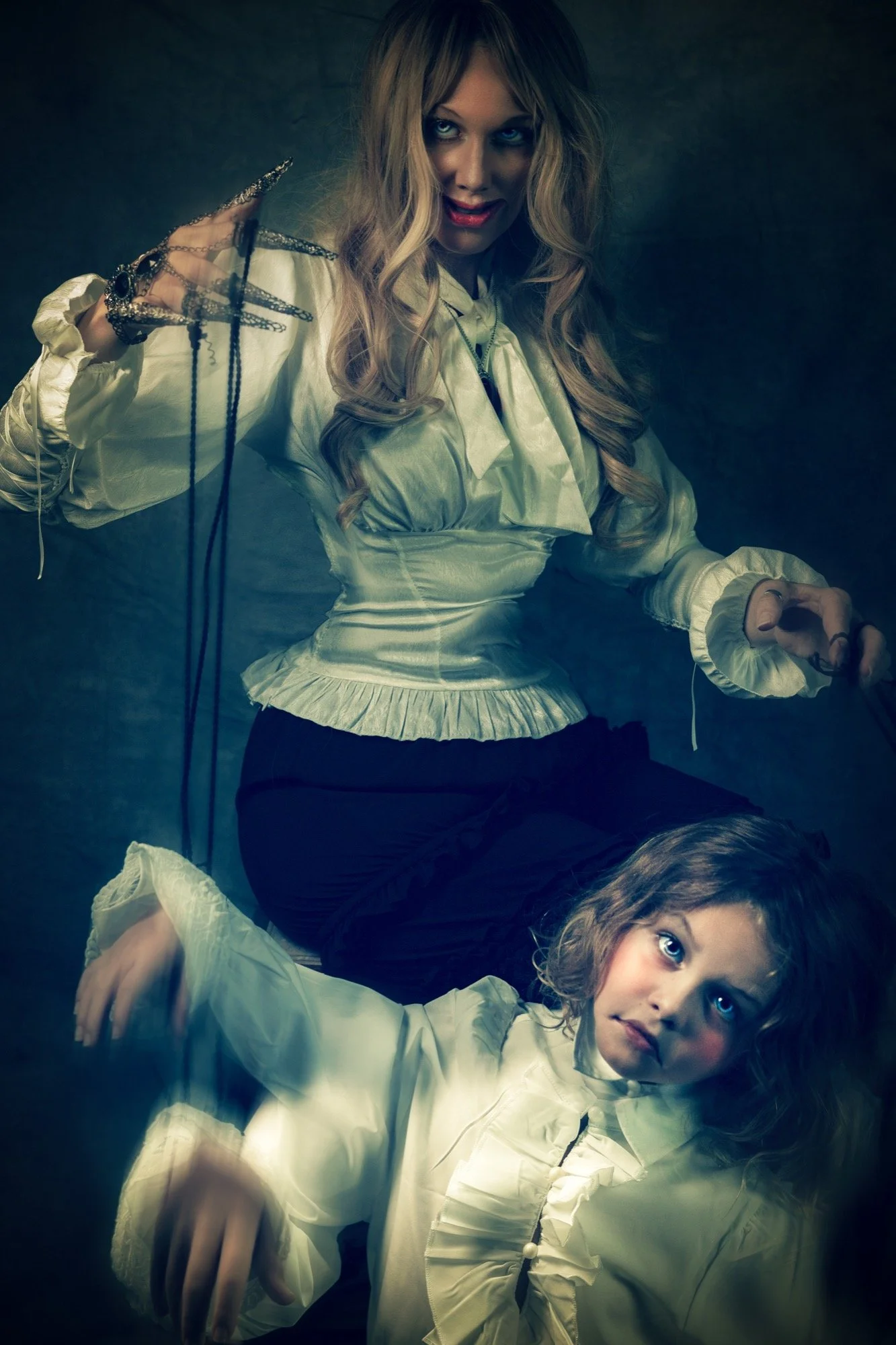 A woman with long, curly blonde hair and a young girl with curly brown hair, both lying on the floor wearing old-fashioned, ruffled, cream-colored blouses with bows. The woman has a mischievous expression and is holding strings attached to the girl, 