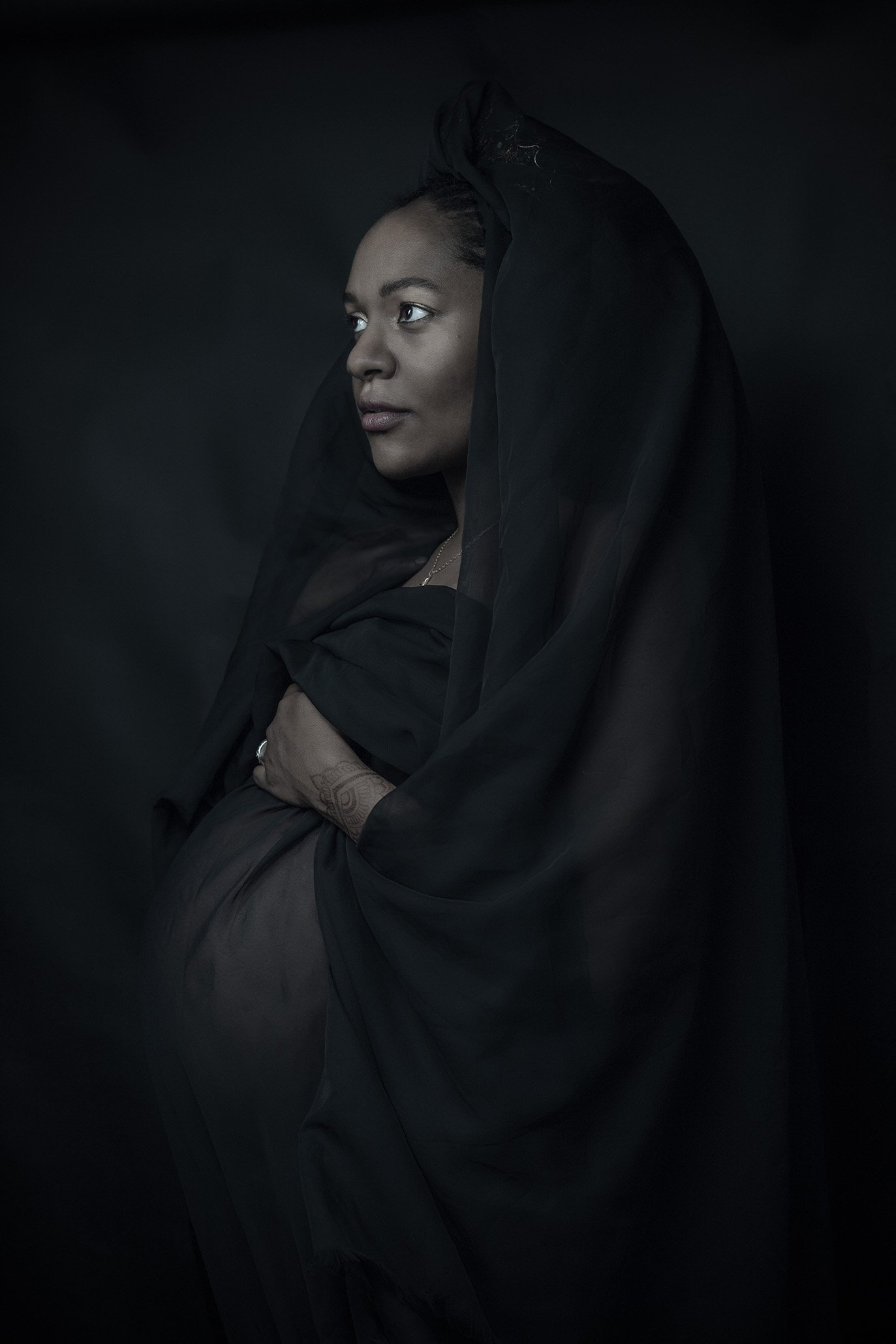 Portrait of a woman in black clothing, softly lit against a dark background, with her head slightly turned to the side.