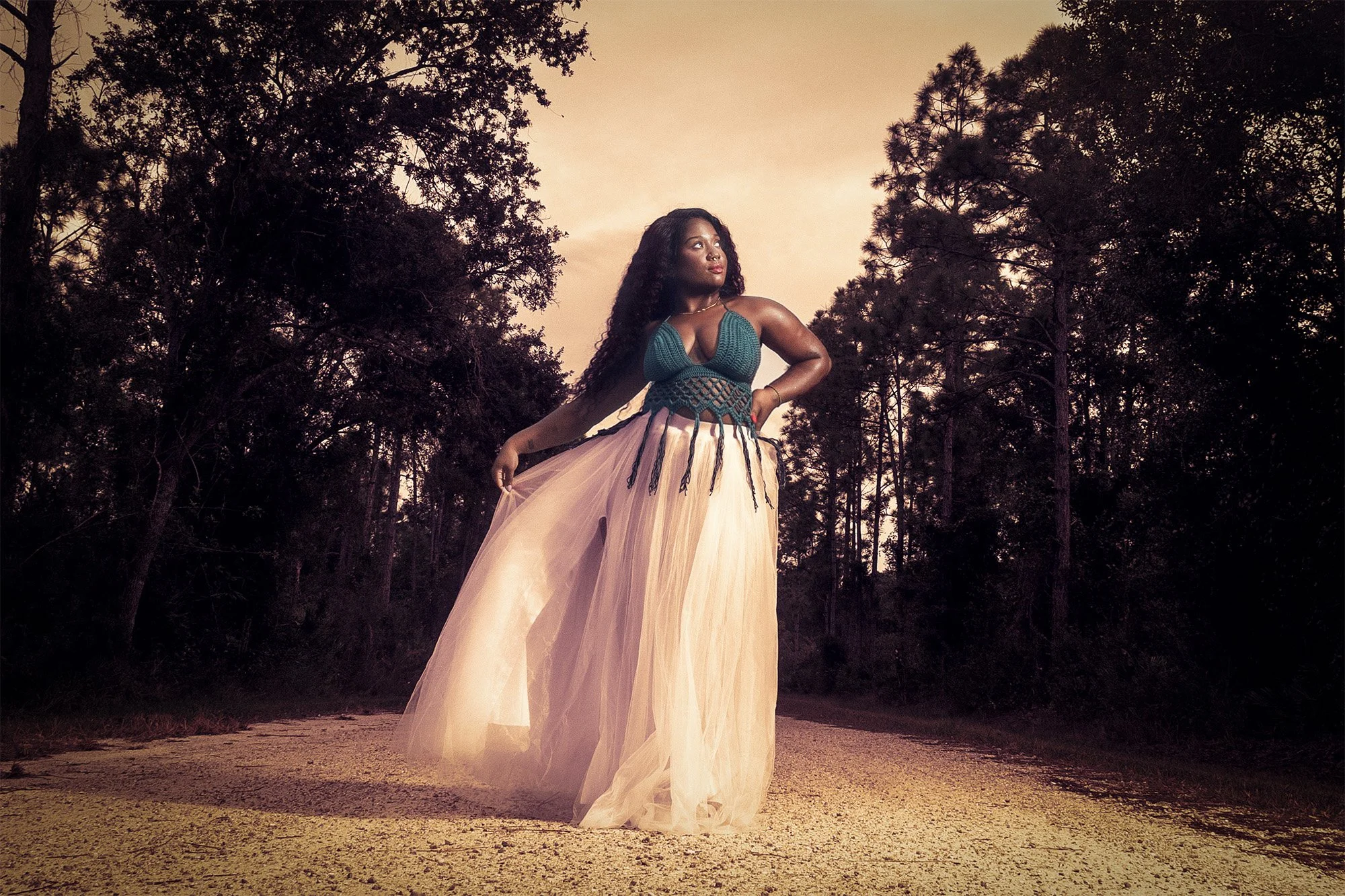 A woman in a flowing peach-colored tulle skirt and a blue crocheted top stands on a dirt road surrounded by trees during sunset.
