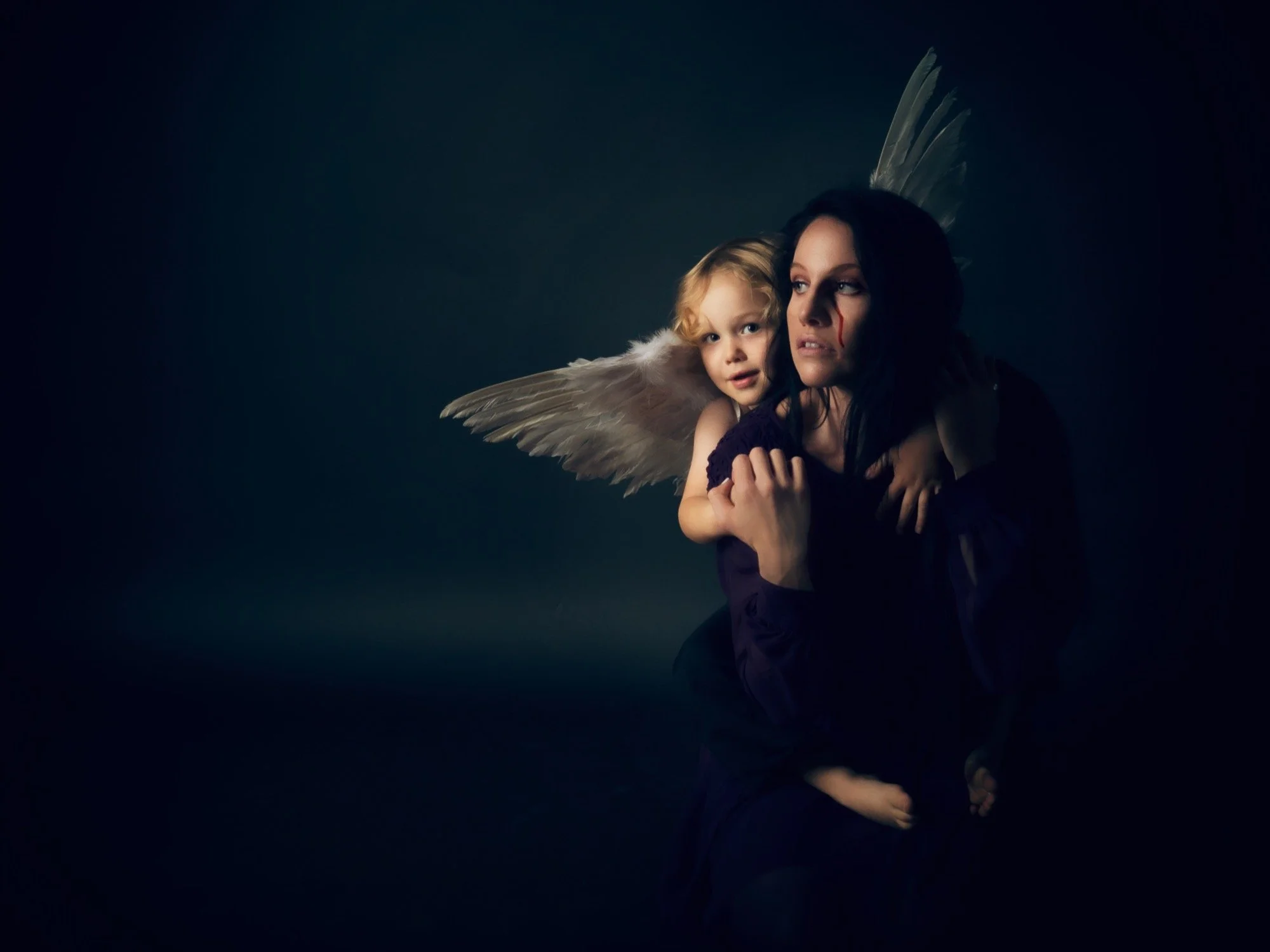 A woman with dark hair and face makeup is holding a young girl with blonde hair and wings on her back. The background is dark with subtle lighting highlighting the subjects.