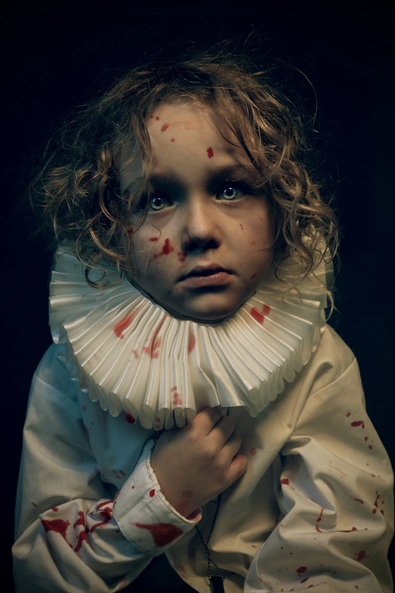 A young child with curly hair and pale skin, wearing a cream-colored outfit with a ruffled collar, has blood stains on their face, hands, and clothing, with a dark background.