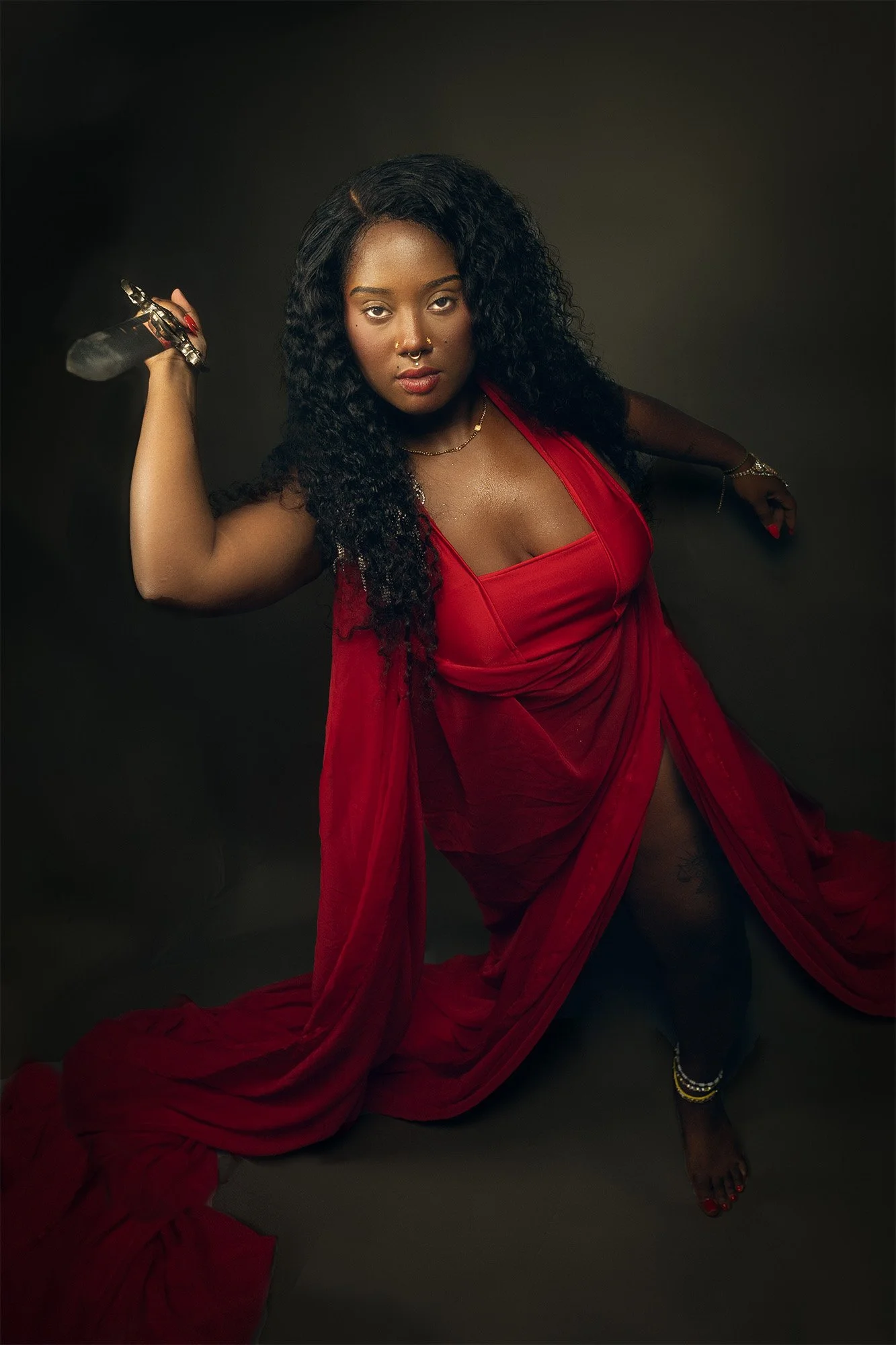 A woman in a red dress holding a sword over her shoulder, looking confidently into the camera against a dark background.