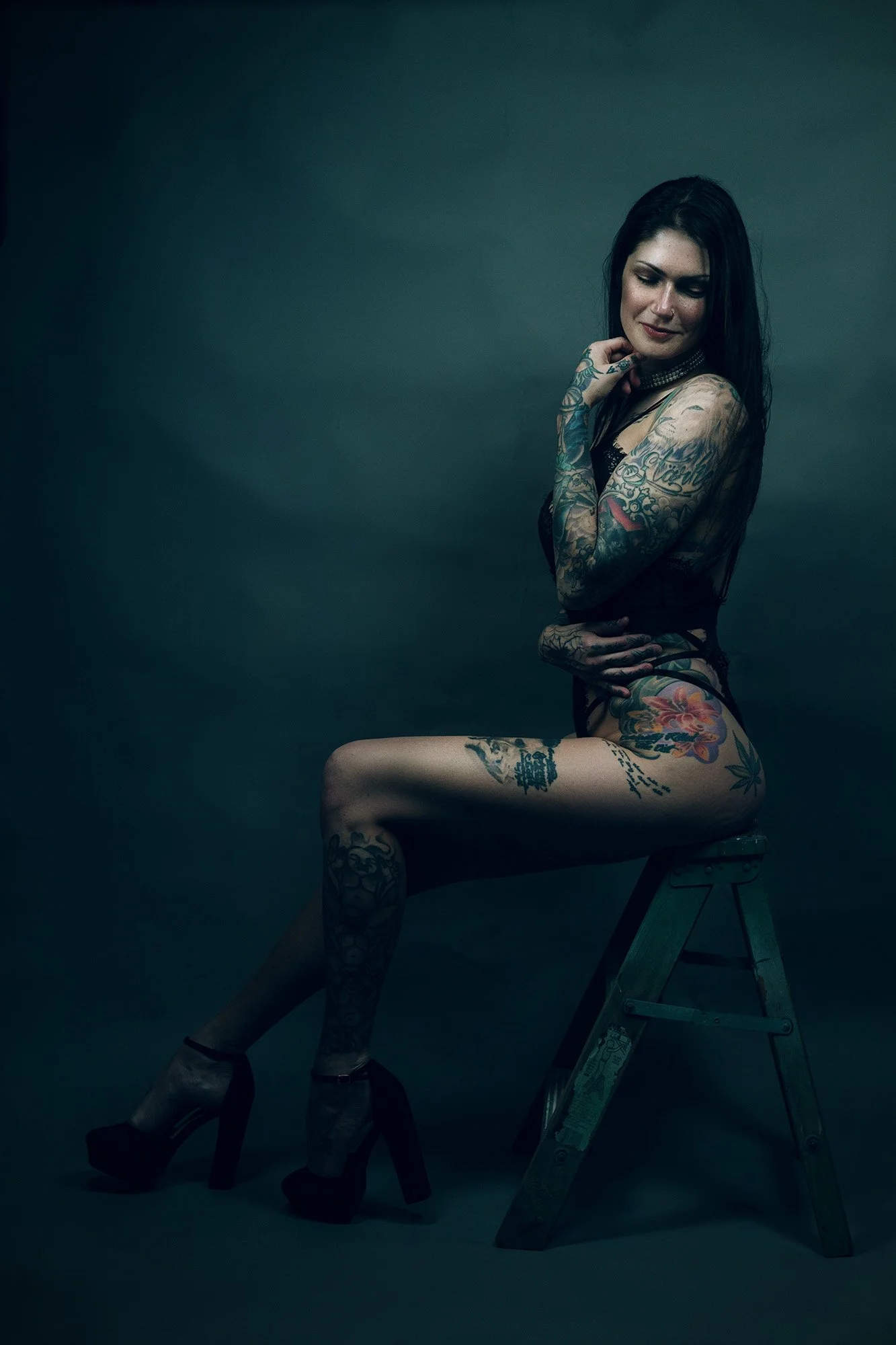 A tattooed woman with dark hair wearing high heels, sitting on a small wooden step ladder against a dark background, with her eyes closed and a slight smile.
