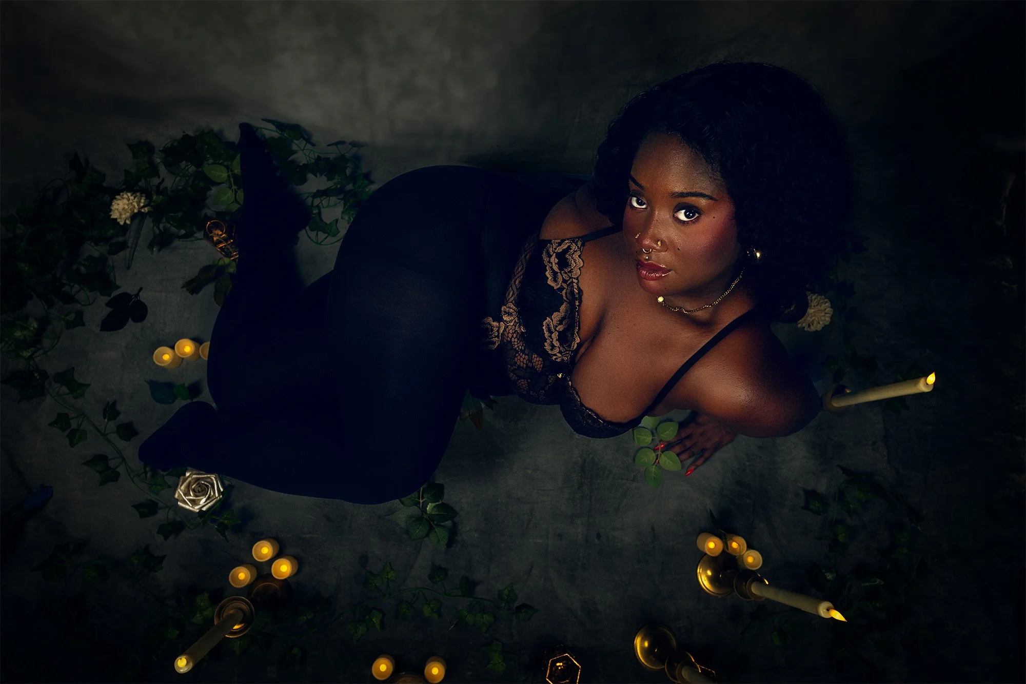 A woman with dark, curly hair and dark skin, dressed in a black dress with lace details, sitting on a dark surface surrounded by lit candles, green foliage, and a silver rose decoration, looking up at the camera with a serious expression.