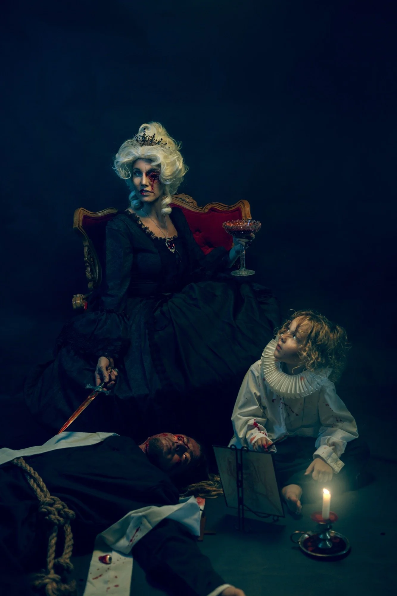 A woman dressed as a queen with white hair, a crown, and black dress, sitting on a red velvet chair with a goblet in her hand. Two children, one with curly hair dressed as a doctor and another lying on the ground with blood on her face, are nearby. S