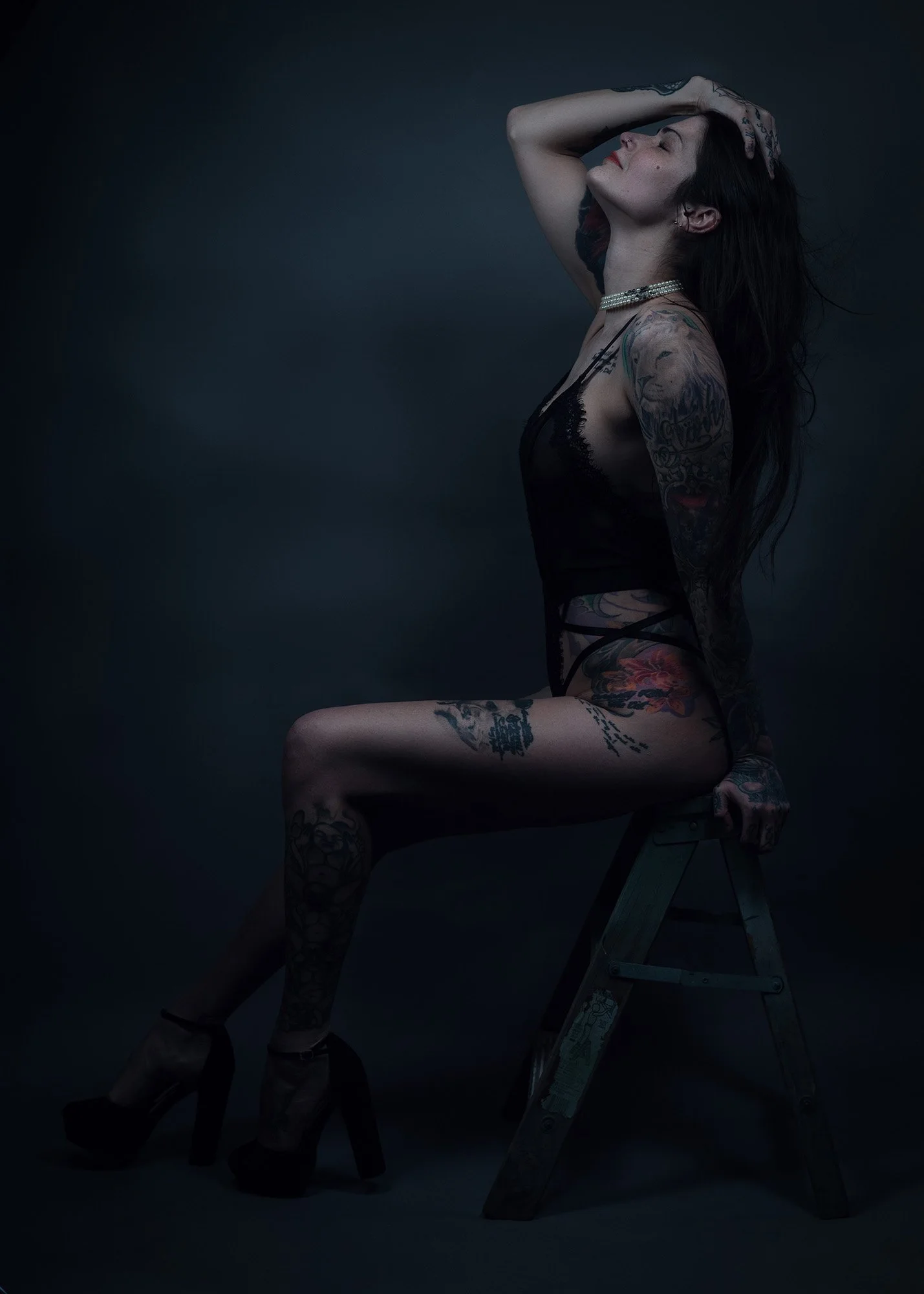 A tattooed woman in black lingerie and high heels sits on a wooden step ladder against a dark background, with one arm raised and her head tilted back.