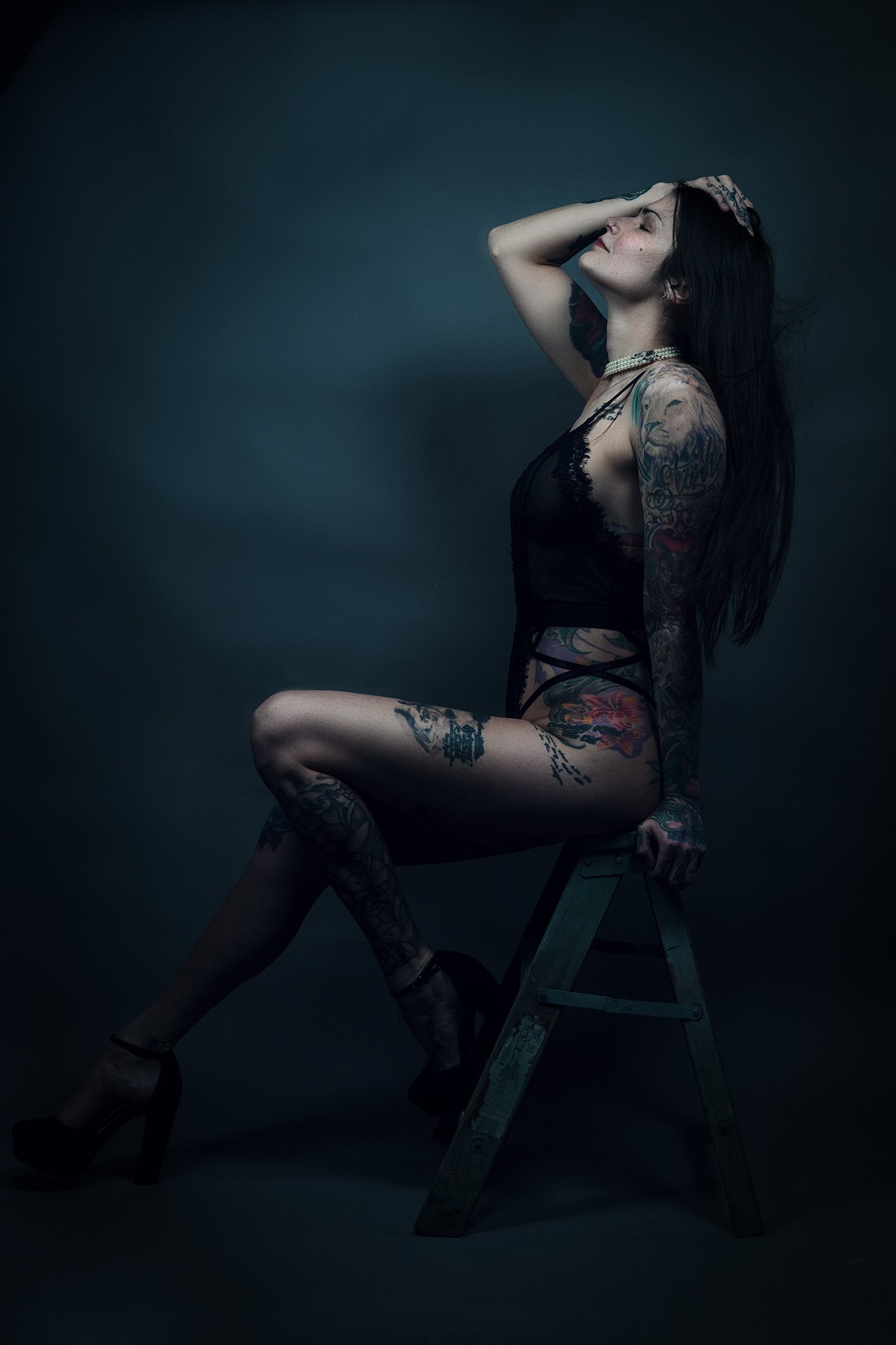 A woman with long black hair and tattoos, sitting on a wooden step ladder, wearing black high heels and a black sheer outfit, with her right hand on her head and eyes closed.