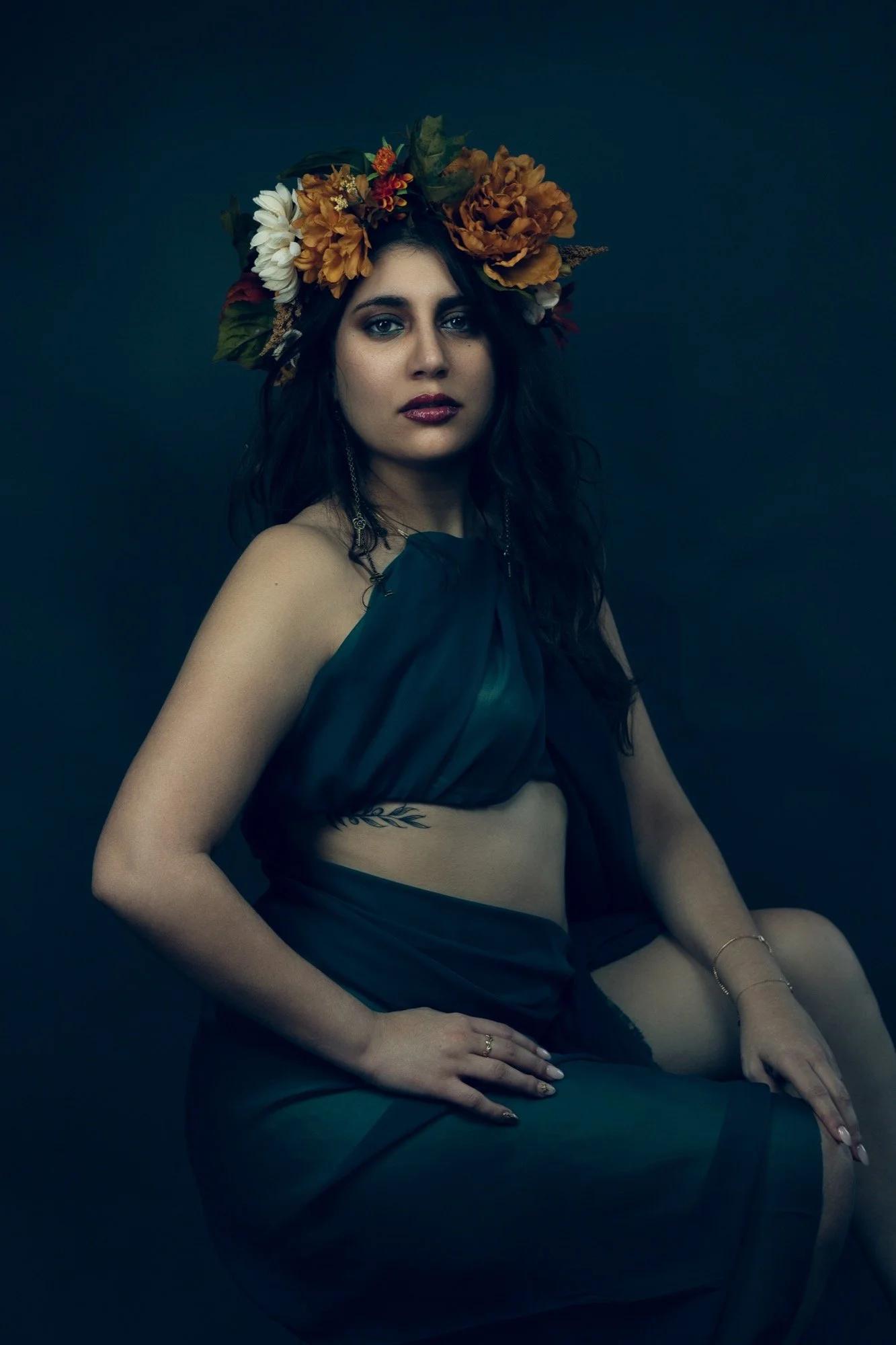 A woman with dark curly hair wearing a large floral crown and a dark, sheer dress sitting against a dark background.