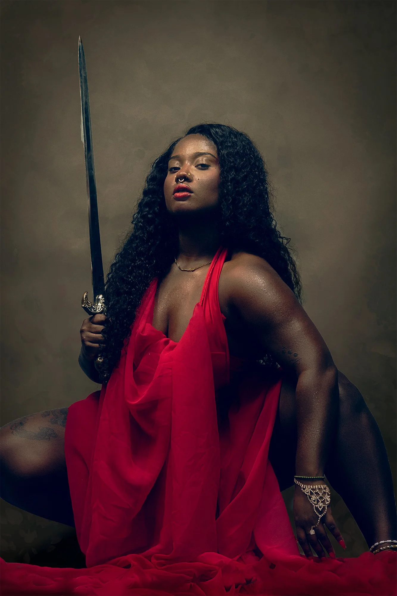 A woman with dark curly hair and dark skin, dressed in a flowing red outfit, holds a sword in her right hand. She is kneeling on a draped red fabric, with a serious expression on her face, against a plain brown background.