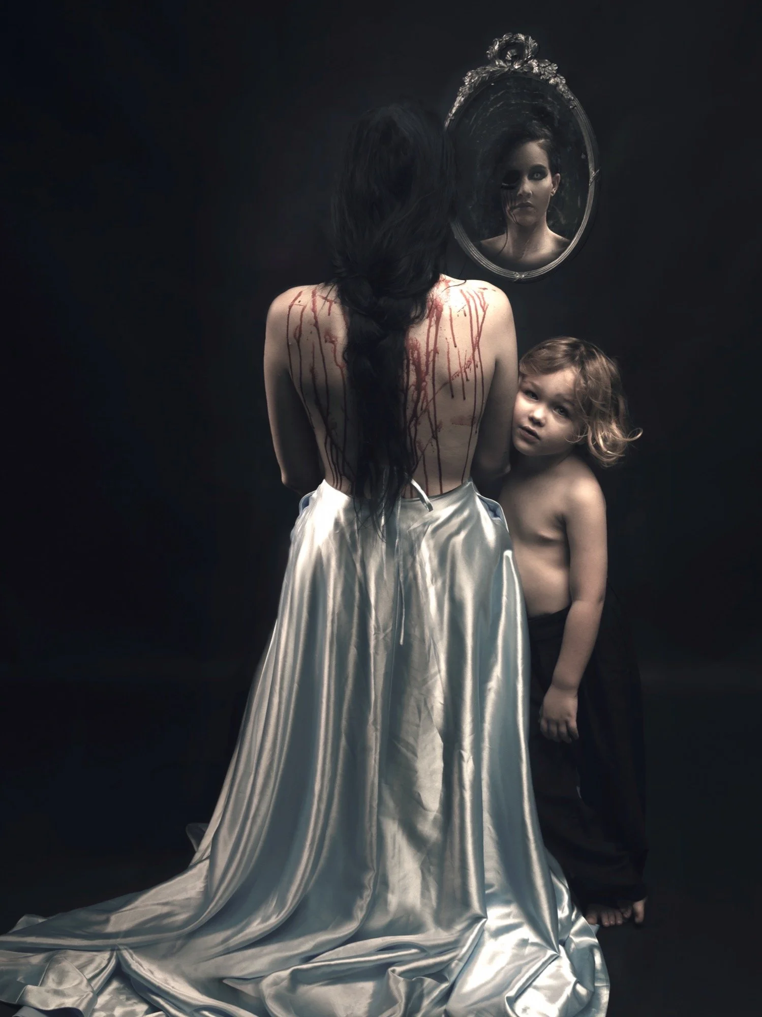 A woman with long dark hair, topless with blood-like streaks on her back, wearing a shiny silver skirt, facing away from the camera, standing in front of a mirror. A young boy with curly blond hair, shirtless, wearing black pants, stands beside her, 