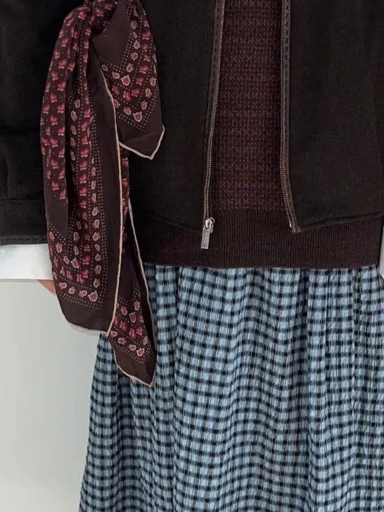 Close-up of a person wearing a black jacket, a dark red zip-up sweater, a brown scarf with pink patterns, and a blue checkered skirt.