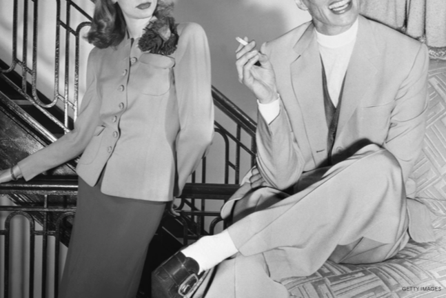 A black-and-white photo of a woman in a blazer and skirt standing next to a man sitting on a couch, holding a cigarette, both dressed in vintage clothing, with a staircase in the background.