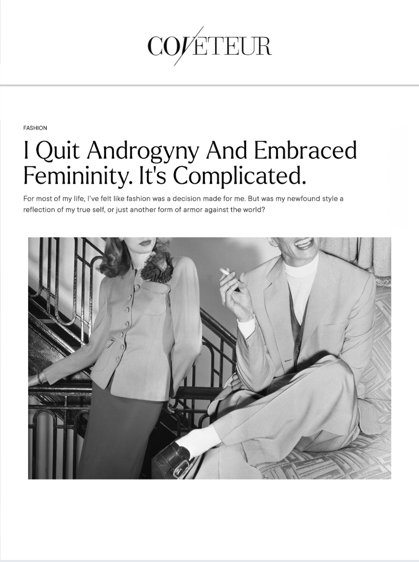 A black-and-white photo of a woman in a blazer and skirt standing next to a man sitting on a couch, holding a cigarette, both dressed in vintage clothing, with a staircase in the background.