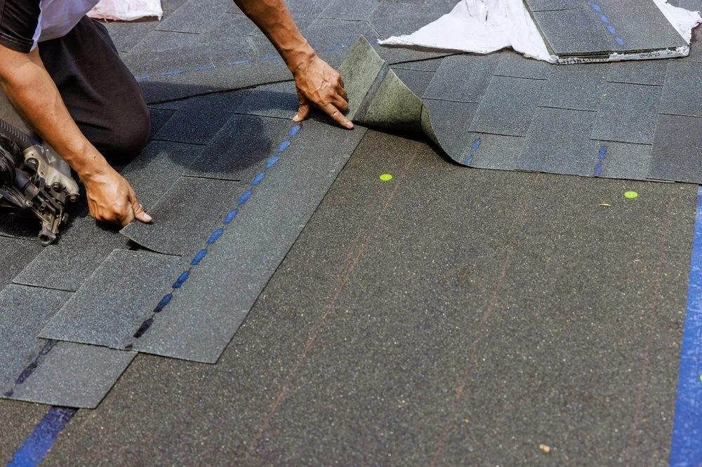 Person installing asphalt shingles on a roof, laying shingles in a row with blue chalk guides.