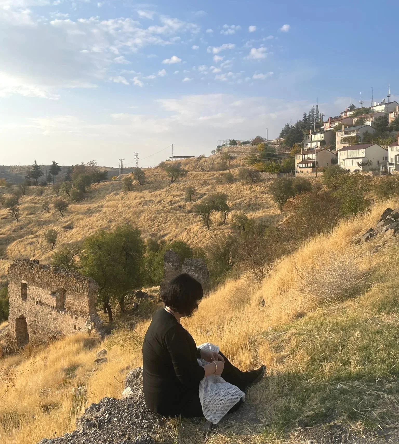 Deborah crocheting Satinig's story overlooking the remains of Sourp Hagop on the eastern side of Kharpert, Türkiye; beyond are the hills where the Euphrates College once stood, 2025. Photo credit: © Ezgi Kilincaslan 2025.