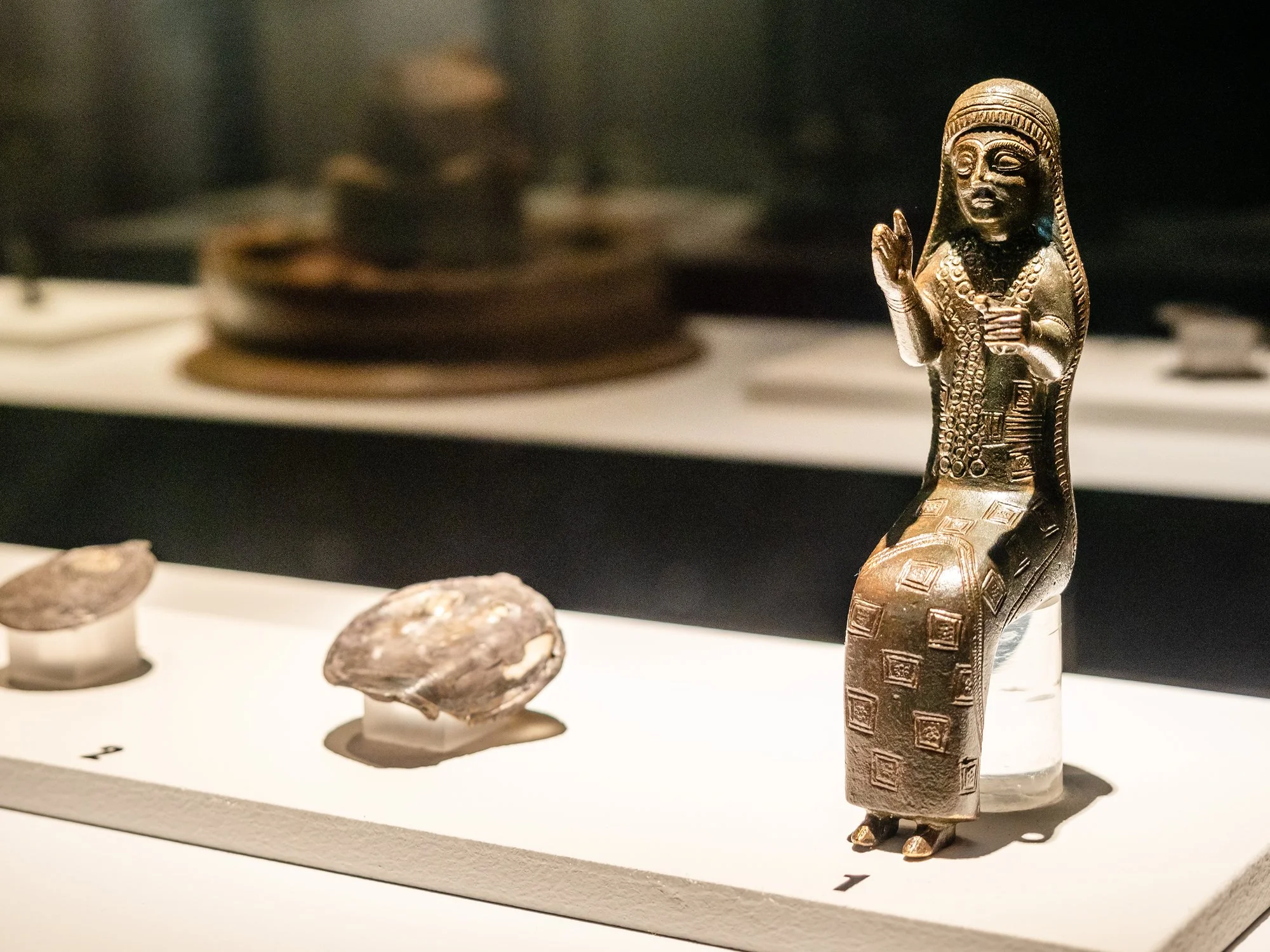 Small, ancient, dark bronze statue of female-appearing figure with outstretched arms and lace-trimmed head covering