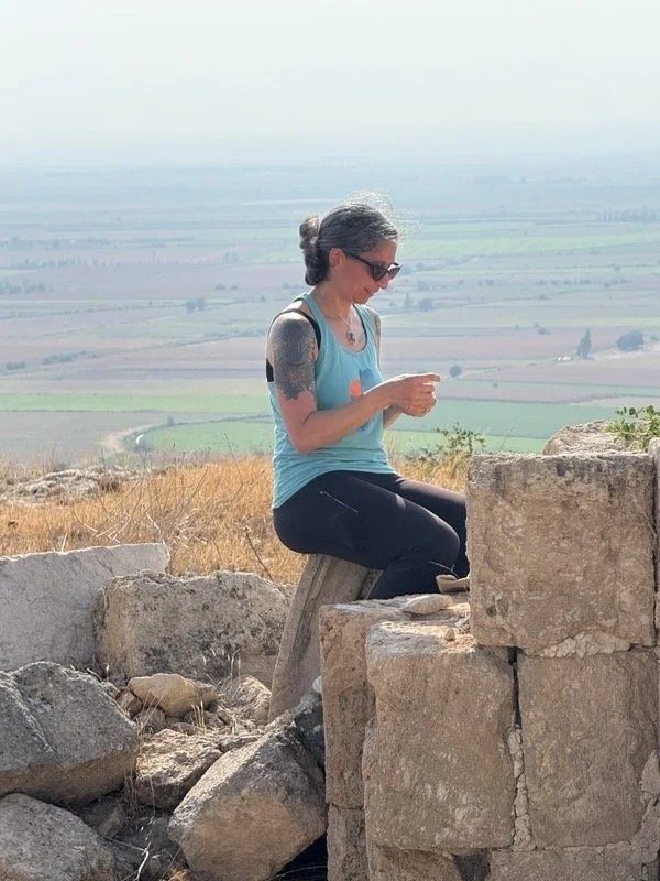 Armenian Needlelace Initiative co-founder Elise Youssoufian sitting on ancient stone ruins and making Armenian needlelace, with green valley farmland in background