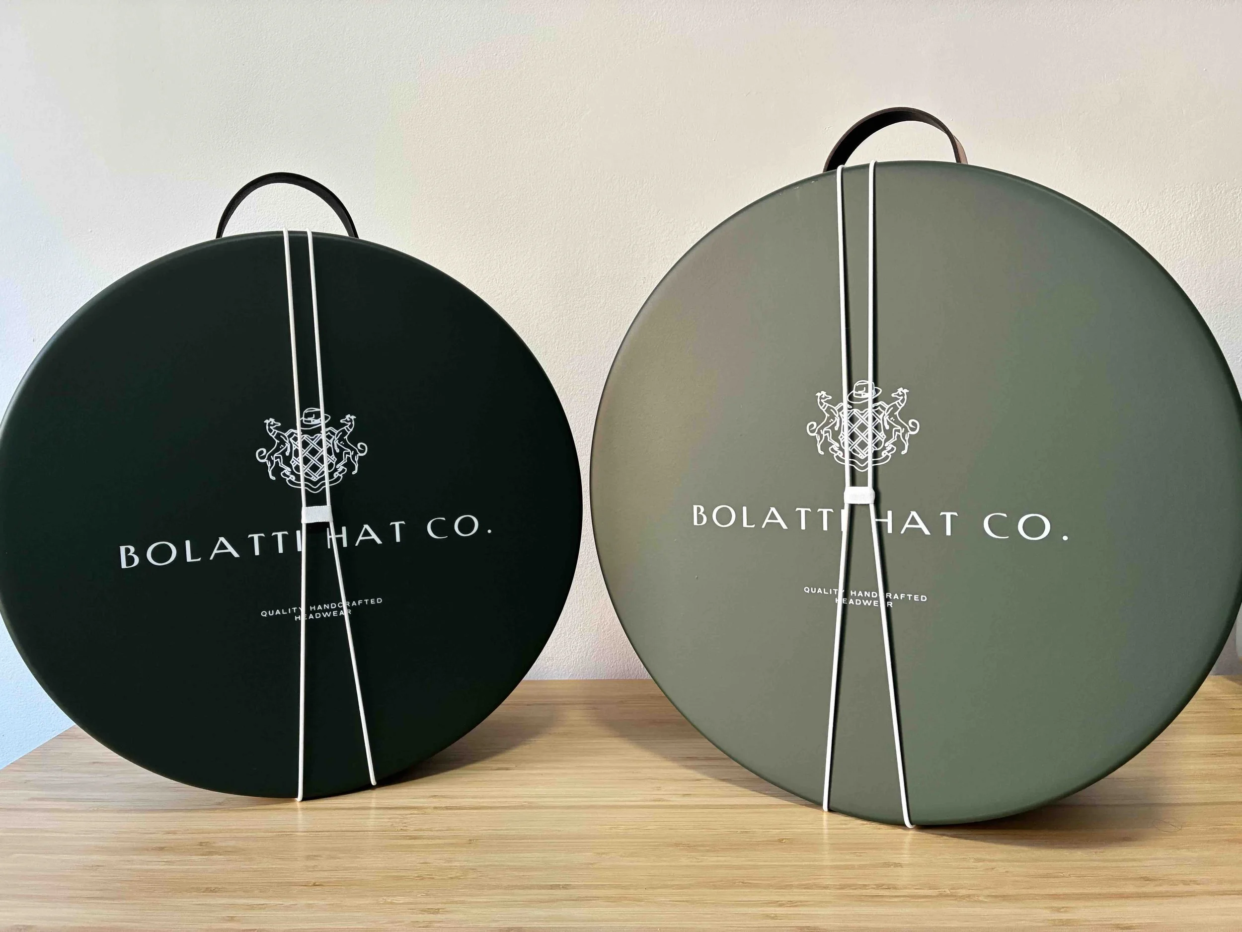 Dos cajas redondas de color verde oscuro con correas blancas, etiquetadas con 'BOLATTI HAT CO.' y un logotipo de escudo, colocadas sobre una superficie de madera frente a una pared blanca.