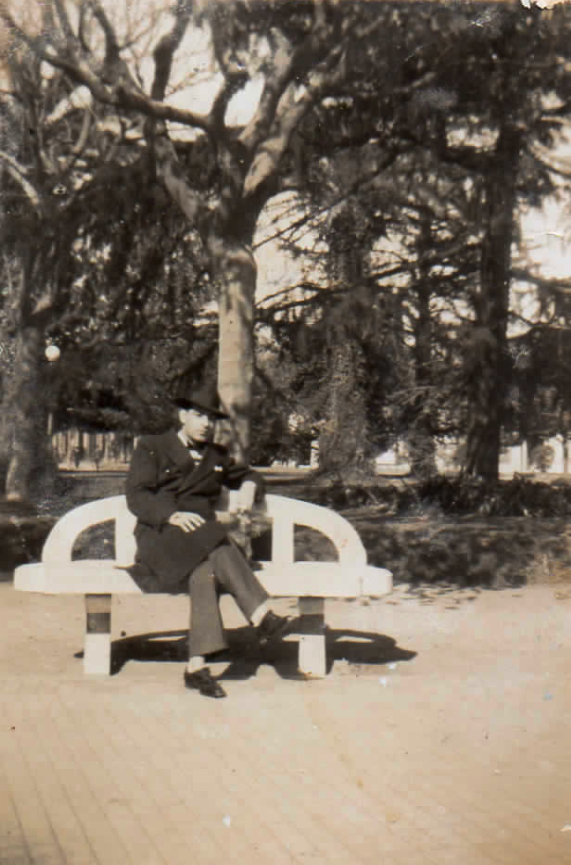 A sepia picture of a finely dressed man with a dark fedora sitting on a curved bench in a park with trees in the background.